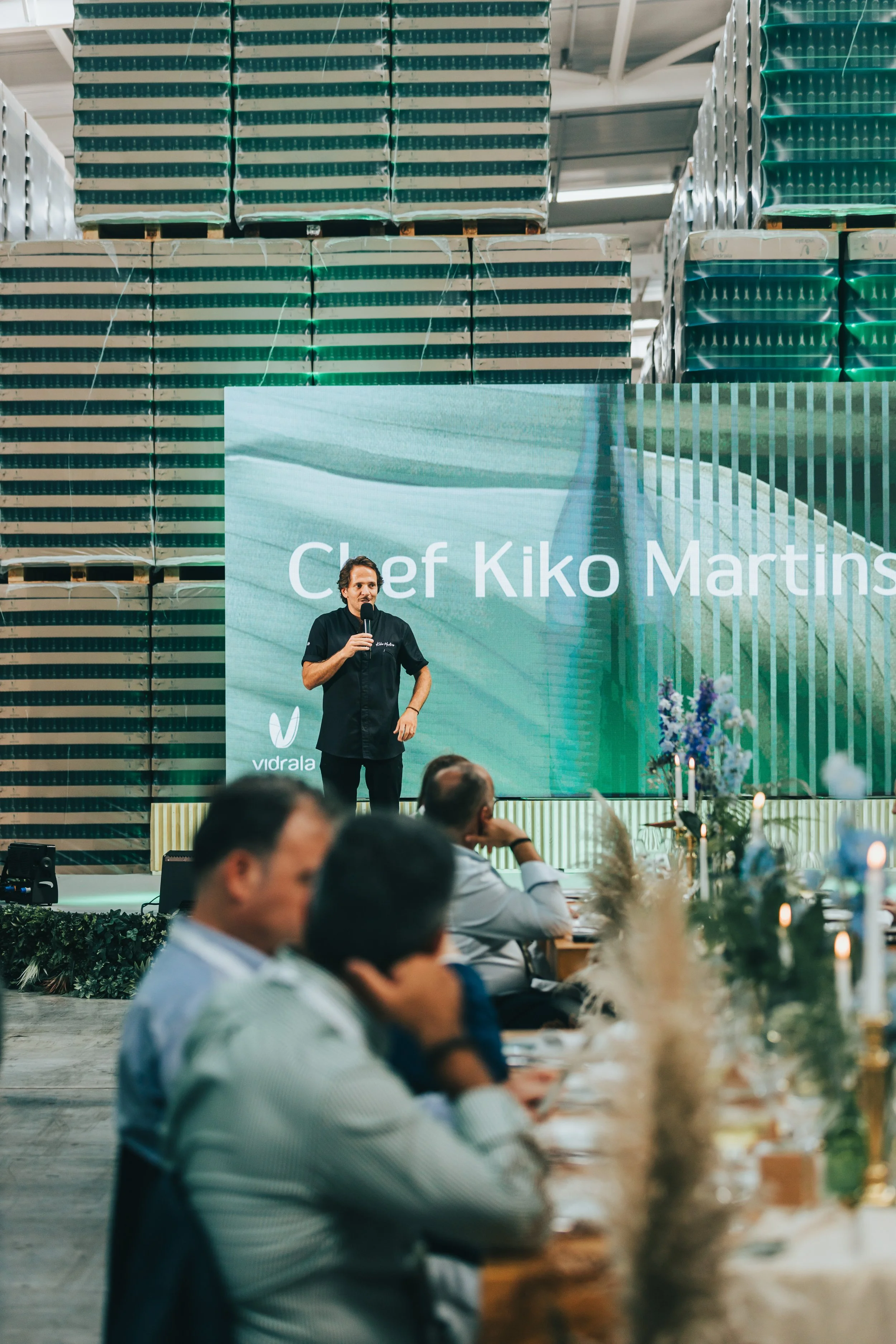Chef Kiko Martins speaking at a conference with a large digital screen behind him displaying his name, in front of an audience at a formal dining event.