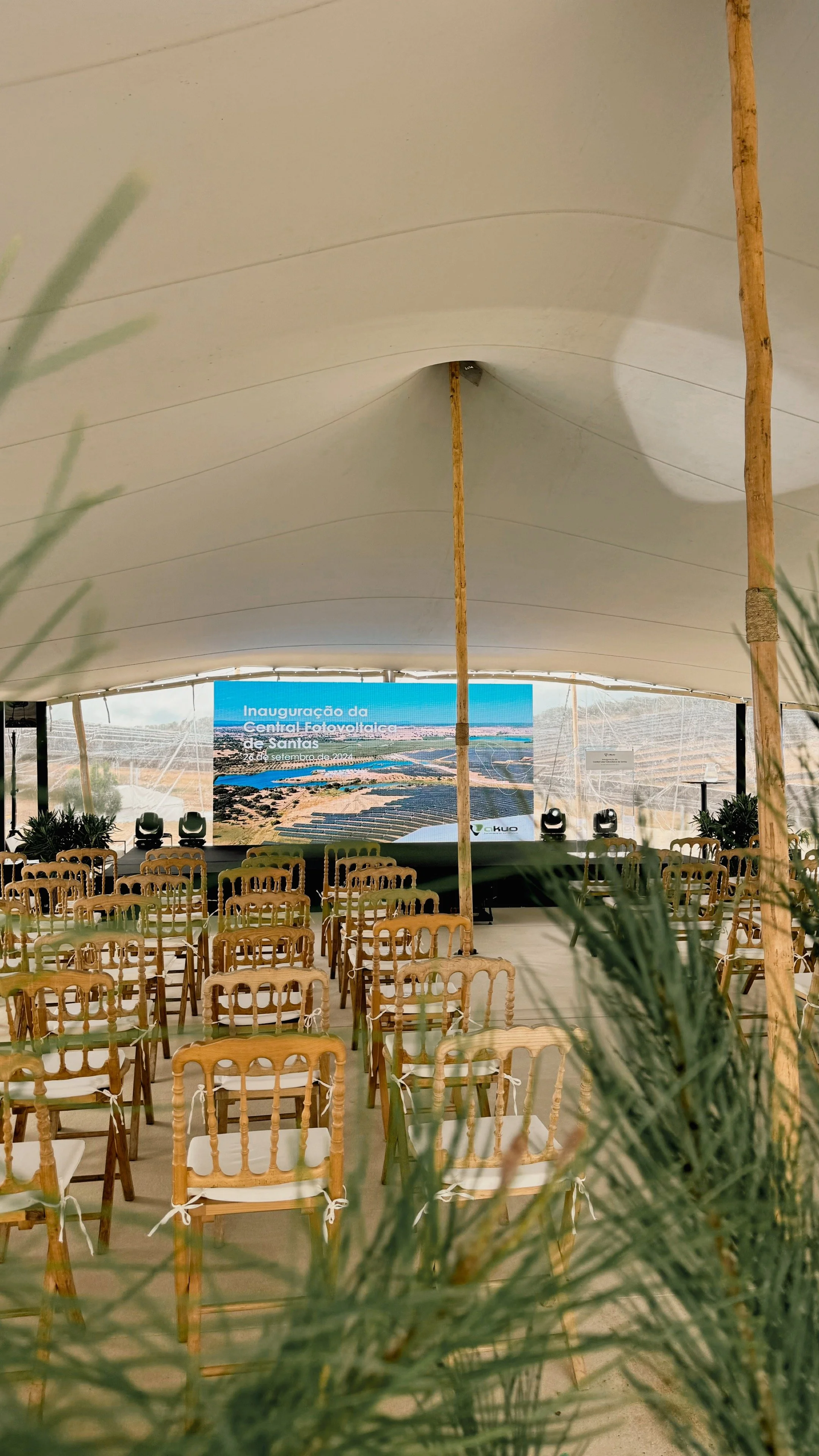 Event space with rows of wooden chairs facing a large screen displaying an aerial view of a solar power facility and a blue sky. The event is for the inauguration of the Central Foto Voltaico de Santa. Wooden support structures hold up a white tent c
