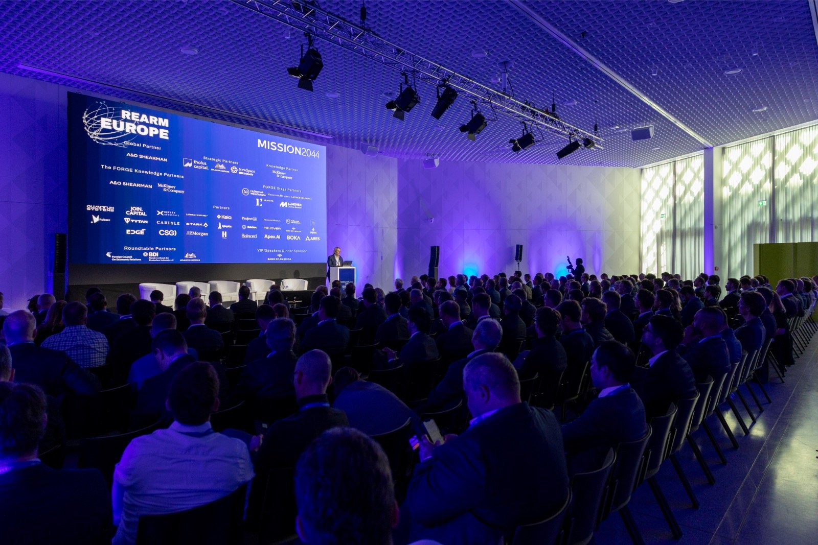 A large conference room filled with people attending a presentation. There is a big screen displaying the 'REARM EUROPE' logo and the title 'MISSION 2044', along with various sponsor logos.