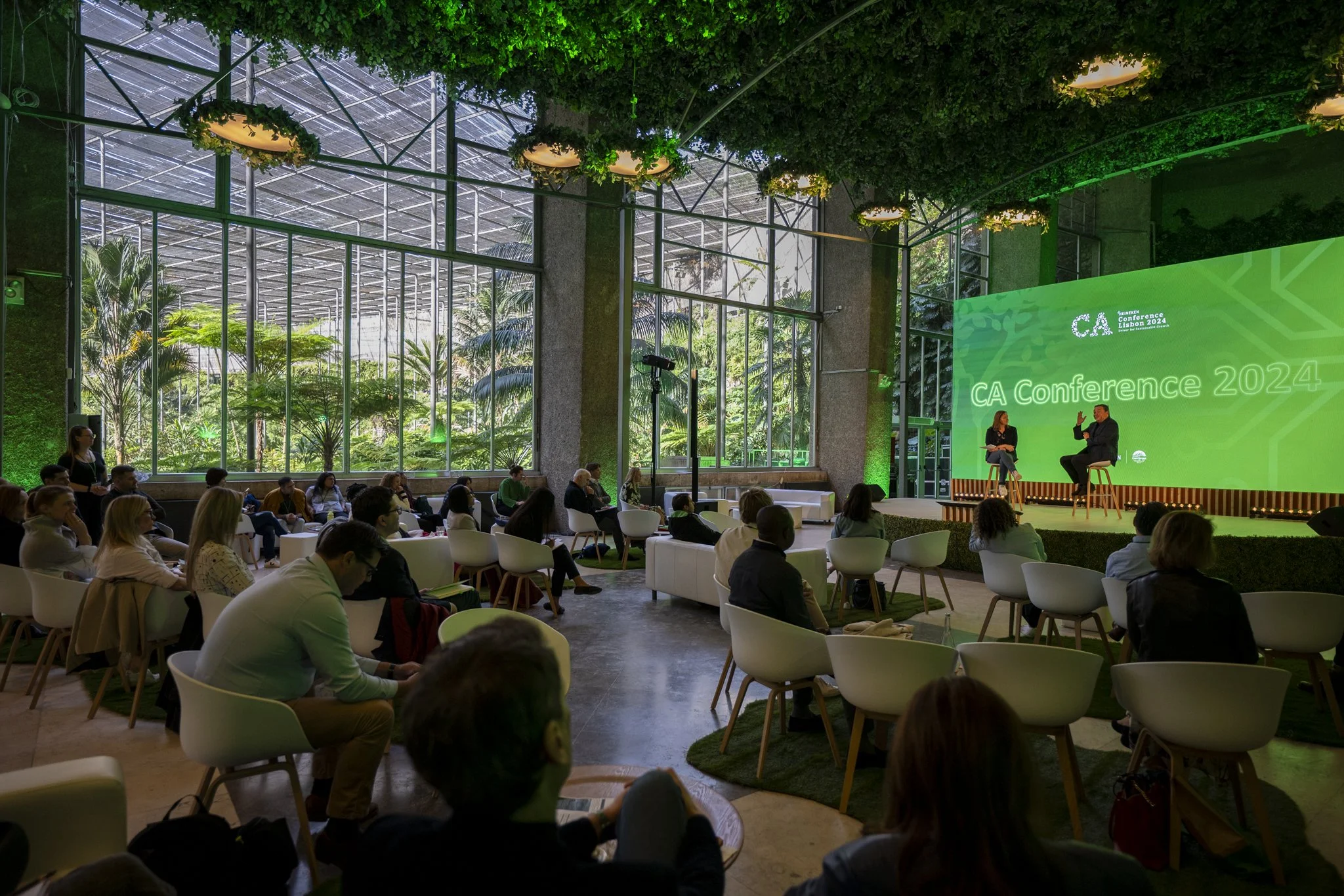 A conference room with large glass windows overlooking lush greenery. Attendees are seated facing a stage where two people are seated and having a discussion. A bright green screen displays conference information.