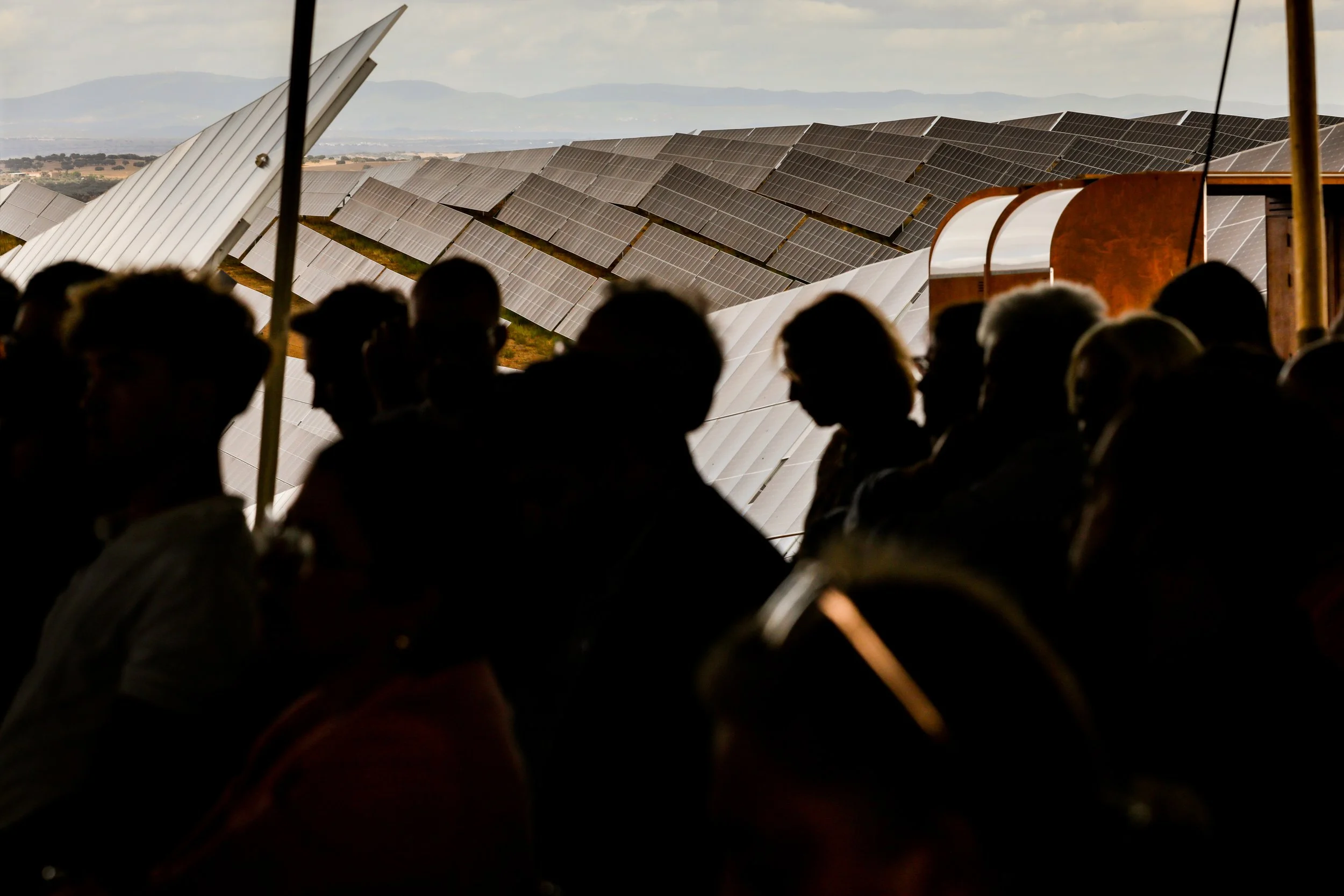 Silhouettes of people seated in front of a large solar panel installation, with mountains in the background.