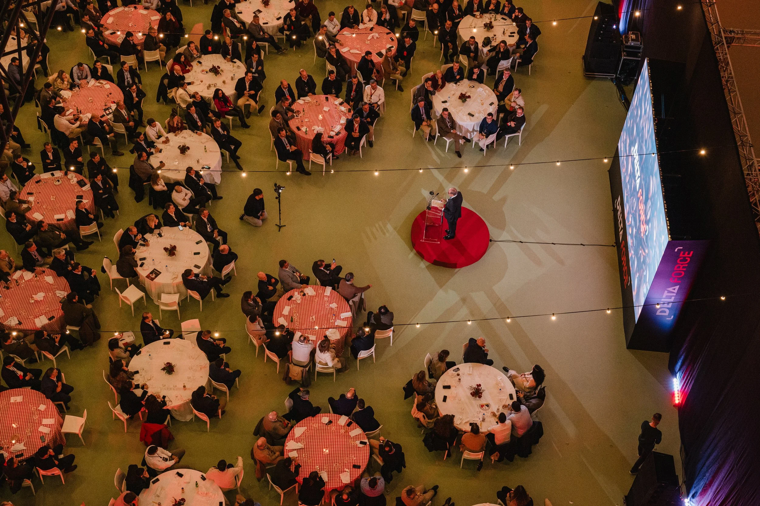 A large indoor event with numerous round banquet tables covered with red and white checkered tablecloths, arranged on green flooring. A speaker in a dark suit stands at a podium with a red carpet, addressing the audience. The audience consists of peo