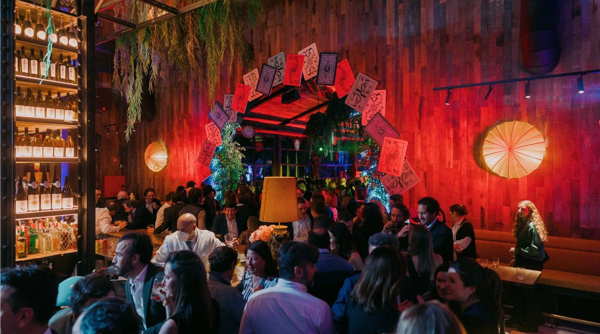 A crowded restaurant or bar with warm lighting, wooden walls, and decorative playing cards hanging above the entrance and walls. People are sitting and socializing at tables and the bar.