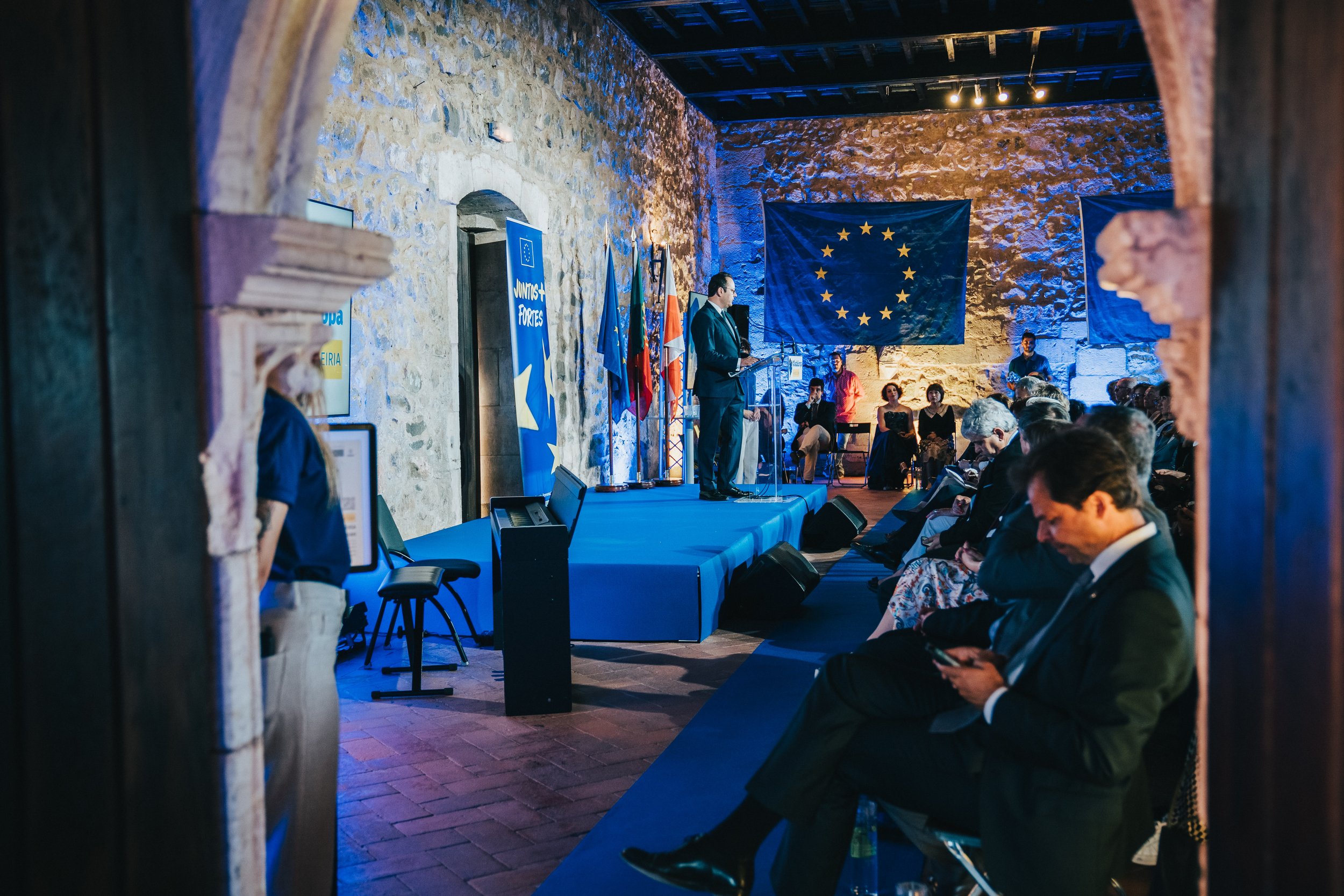 A man is speaking on stage at a conference with European Union flags behind him. The audience appears to be listening attentively in a room with stone walls and blue lighting.