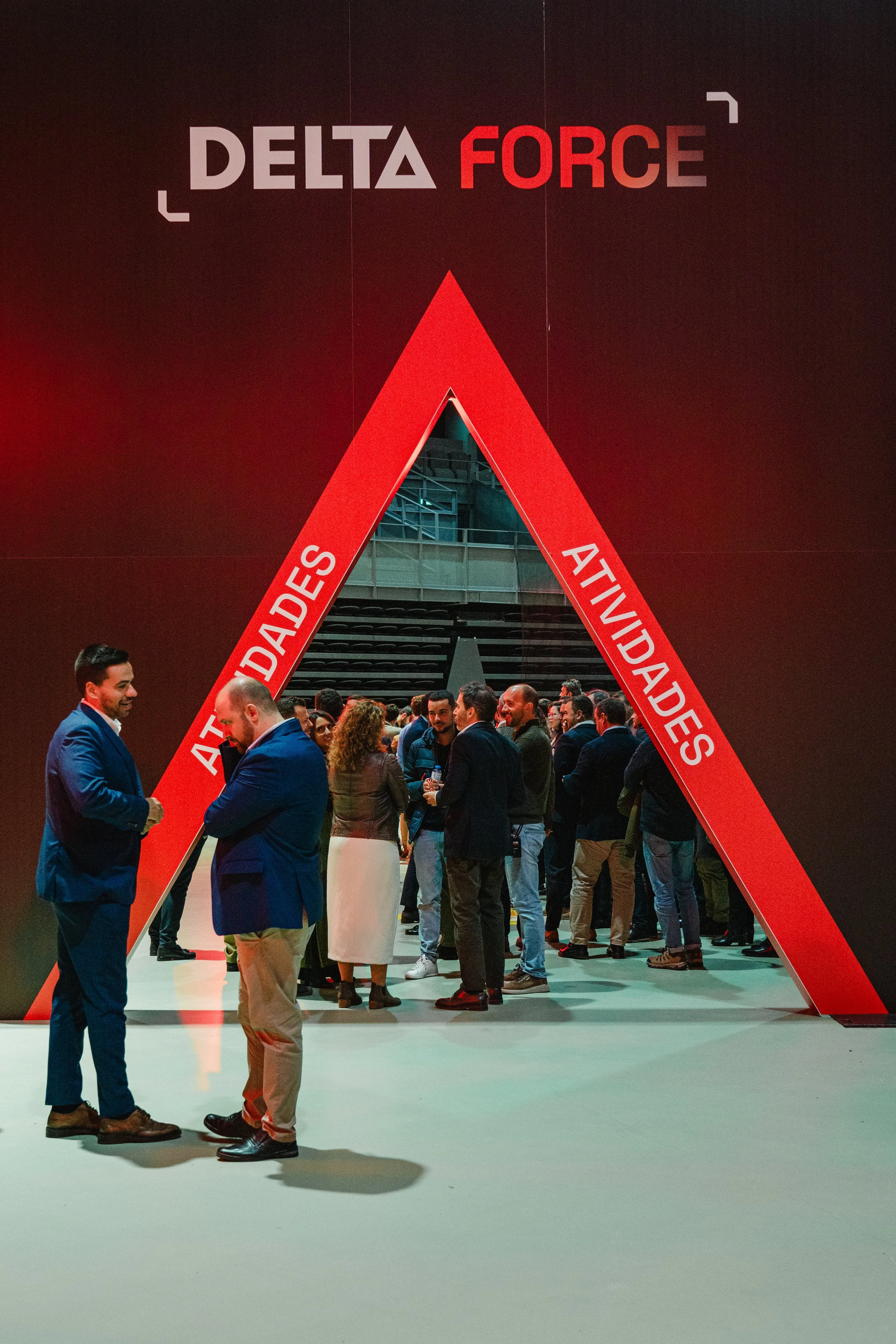 A group of people in business attire gathered at a conference or event, standing and engaging in conversation under a large red triangle structure with the words 'ATADADES' and 'ATIVIDADES' on the sides, and the words 'DELTA FORCE' at the top.