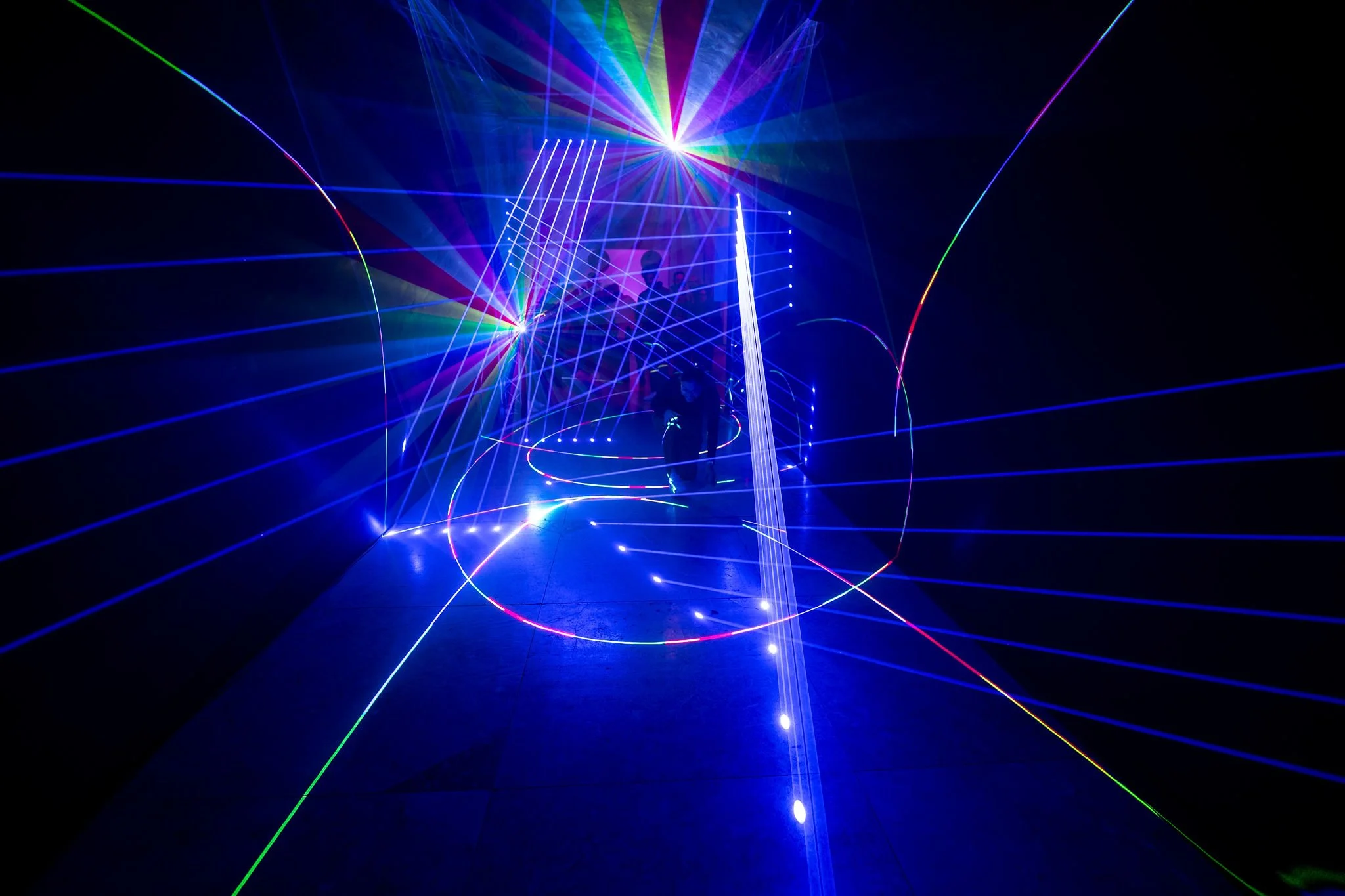 Dark room with vibrant multicolored laser lights creating geometric patterns and beams, with silhouettes of people near a display or stage.