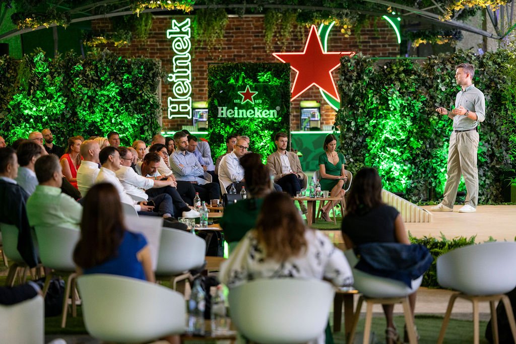 A speaker giving a presentation to an audience seated outdoors at a Heineken event with floral and greenery decor, illuminated Heineken and star signage in the background.