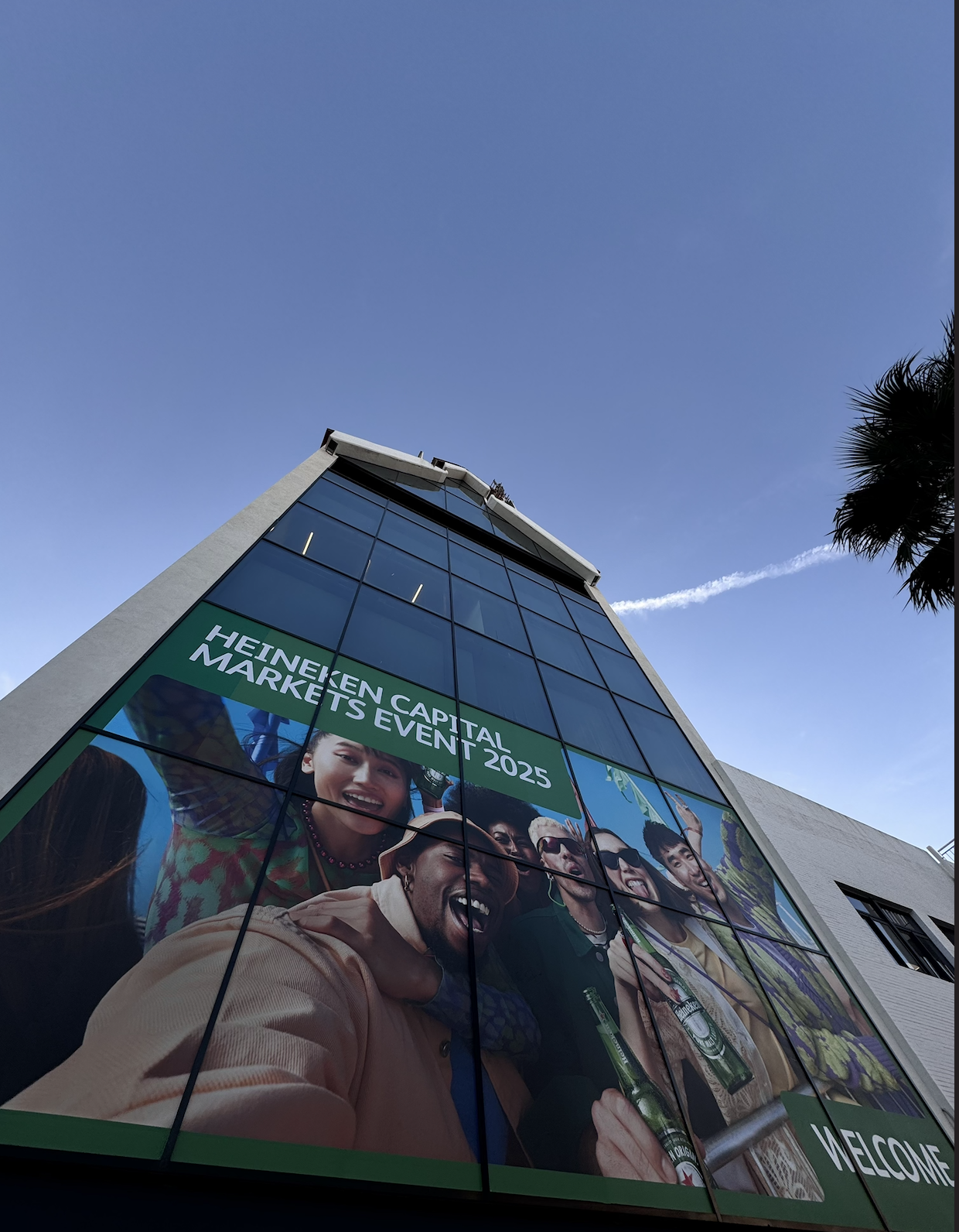 A tall modern building with a large banner displaying people celebrating at the Heineken Capital Markets Event 2025, with bright blue sky and a few clouds in the background.