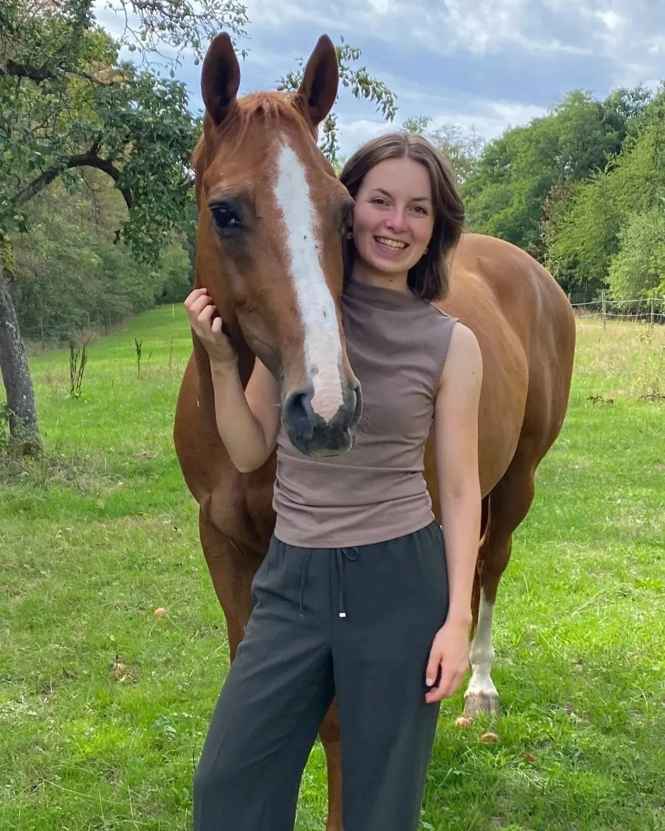 Junge Frau umarmt braunen Pferd auf grüner Wiese mit Bäumen im Hintergrund, bewölkter Himmel.