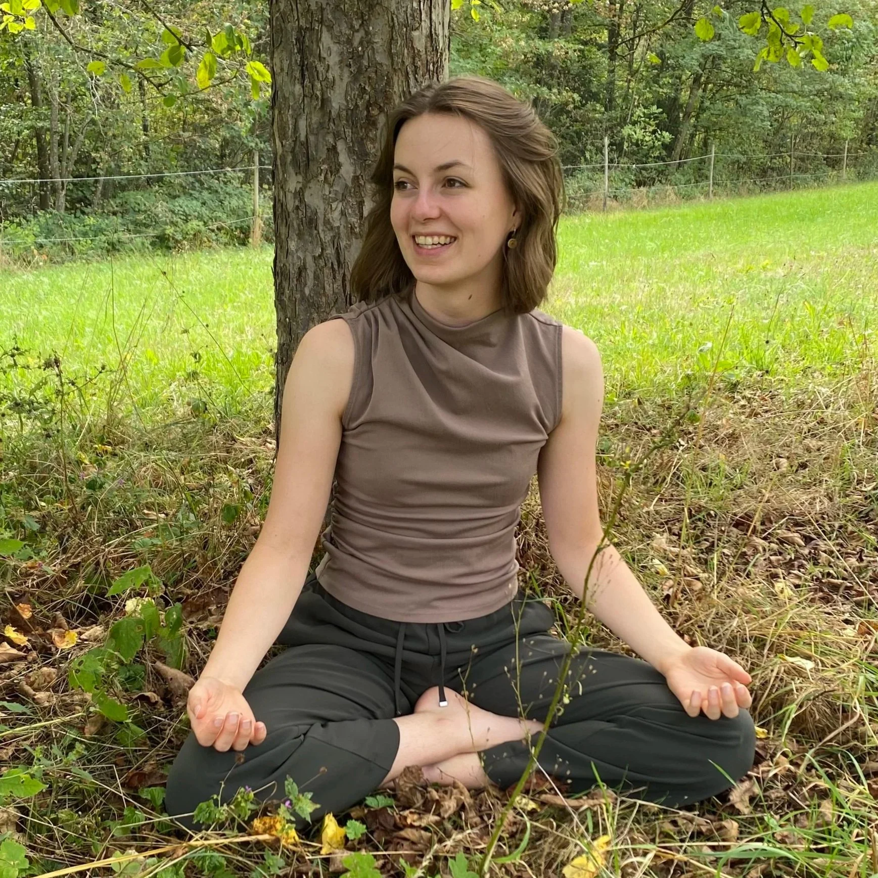 Eine junge Frau sitzt im Freien in der Nähe eines Baumes, macht eine Meditation im Schneidersitz, umgeben von grünem Gras und Bäumen.