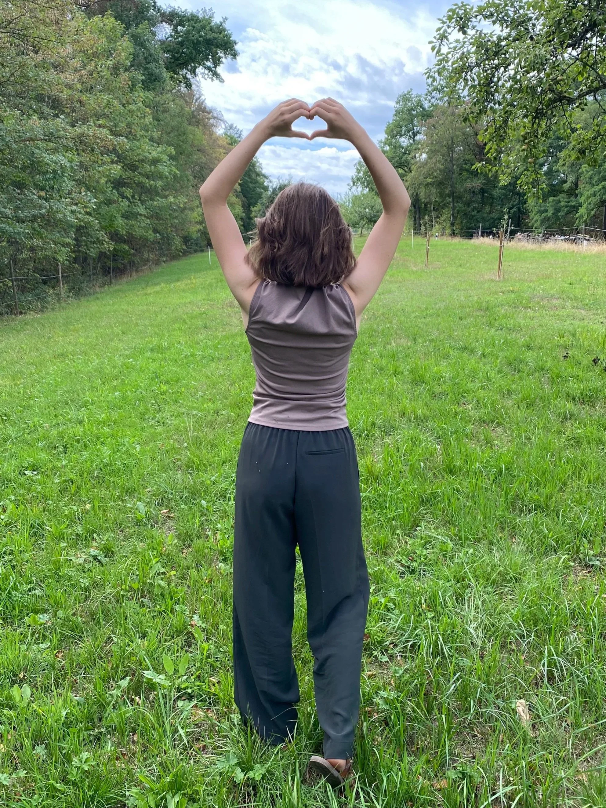 Eine Person steht auf einer Wiese mit Bäumen im Hintergrund und formt mit den Händen in Herzform, Blick nach vorne, bewölkter Himmel.