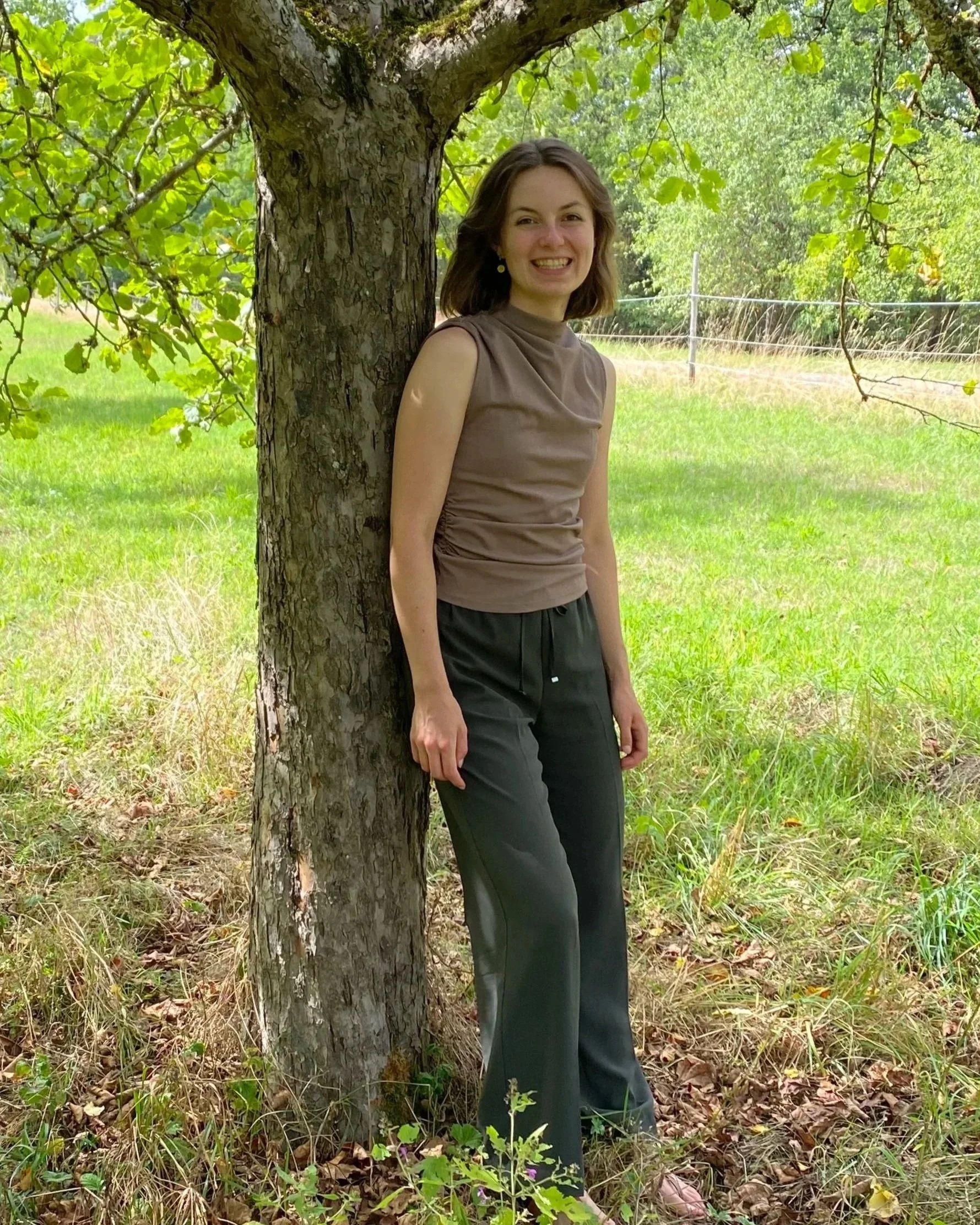 Junge Frau steht mit Rücken an einen Baum im grünen Park, lächelt und trägt ein braunes Top und schwarze Hose.