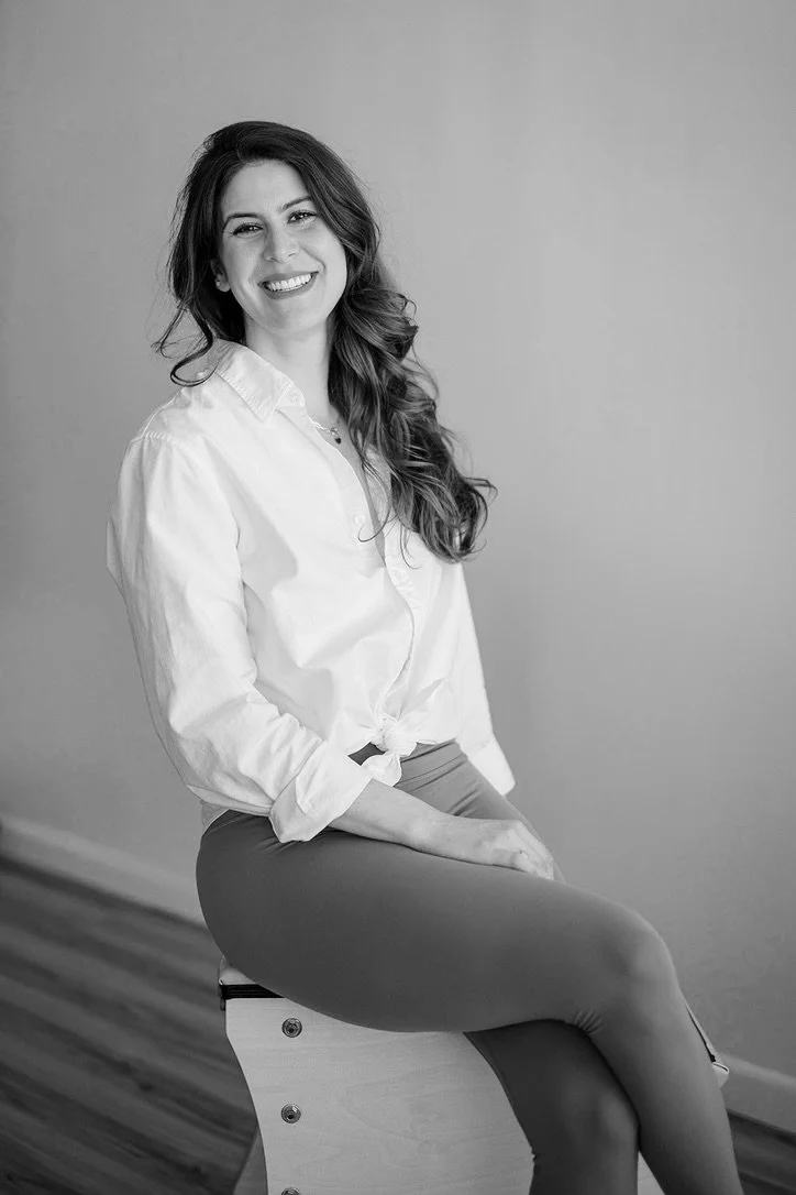 A woman with long, wavy hair sitting on a wooden box, smiling at the camera, wearing a white button-up shirt tied at the waist and dark pants, in a simple indoor setting.