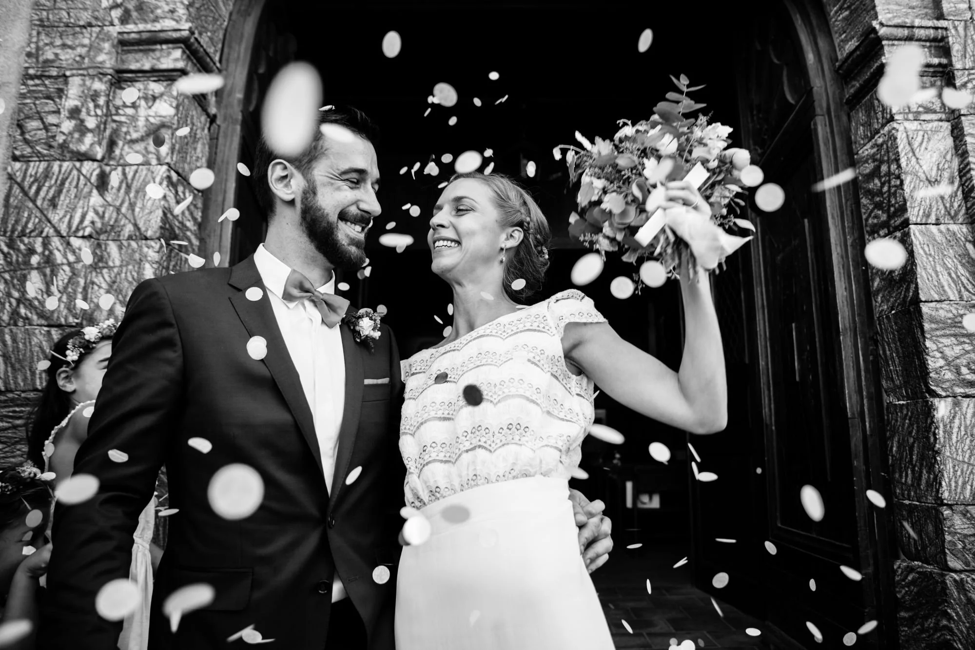 Black-and-white photograph of a bride holding a bouquet beside the groom as confetti falls around them.
