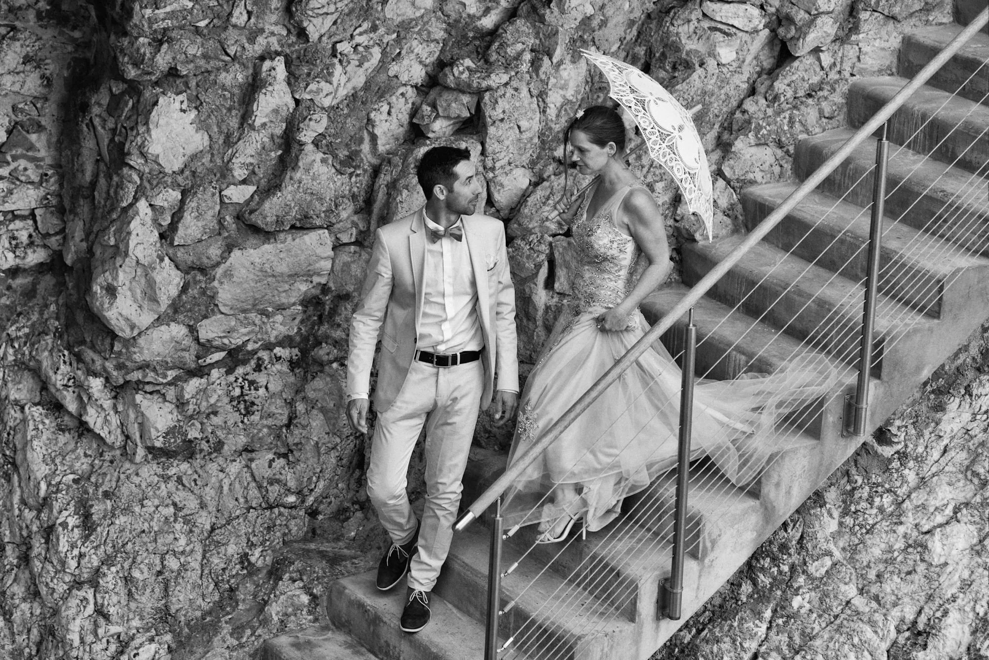 Black-and-white photograph of a bride and groom on steps beside a stone wall.