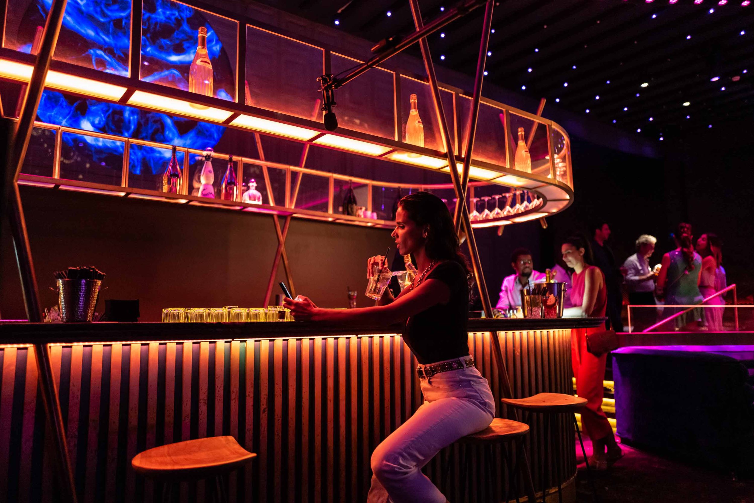 A woman seated at an illuminated bar beneath shelves of bottles and glasses.