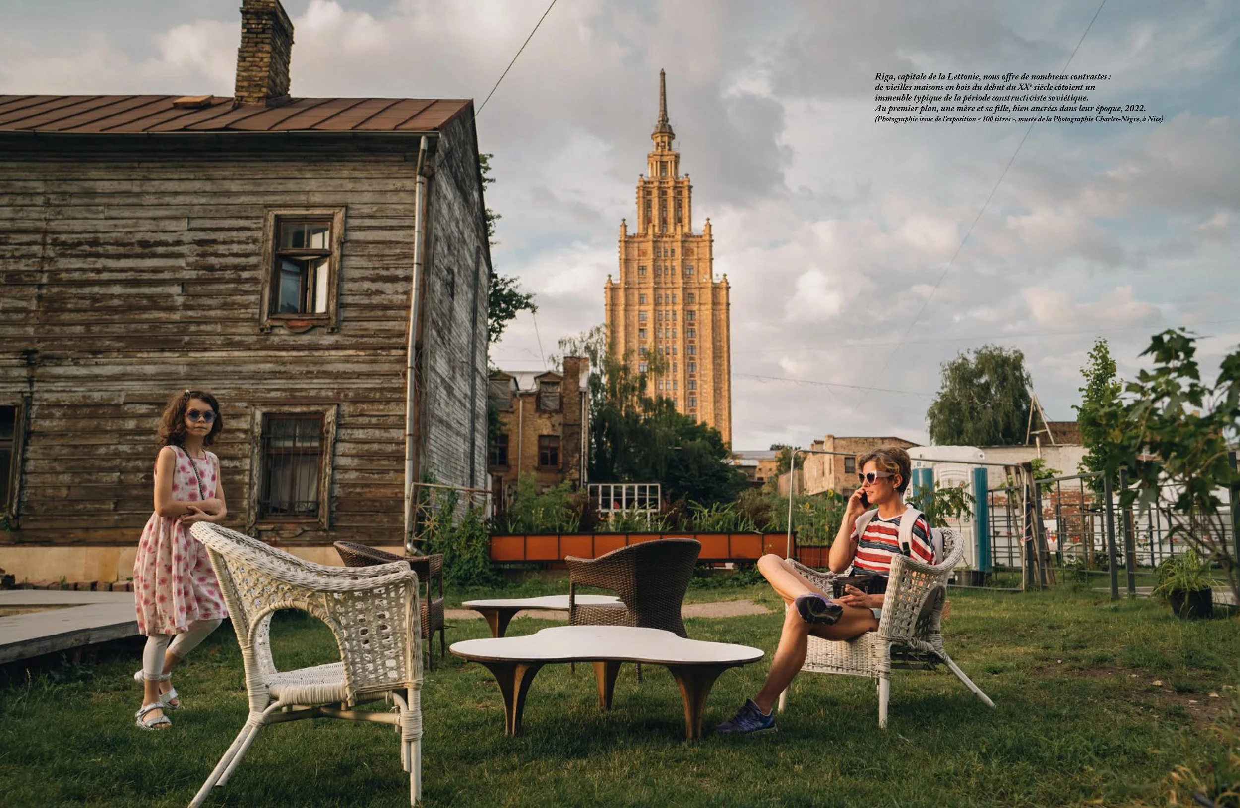 A girl in a pink floral dress standing on a grassy yard, and a person in a striped shirt and sunglasses sitting in a wicker chair on a patio, talking on the phone, with a large historical building in the background, and some trees and garden furnitur