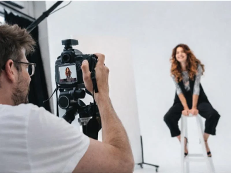A photographer photographing a woman seated on a stool against a white backdrop.