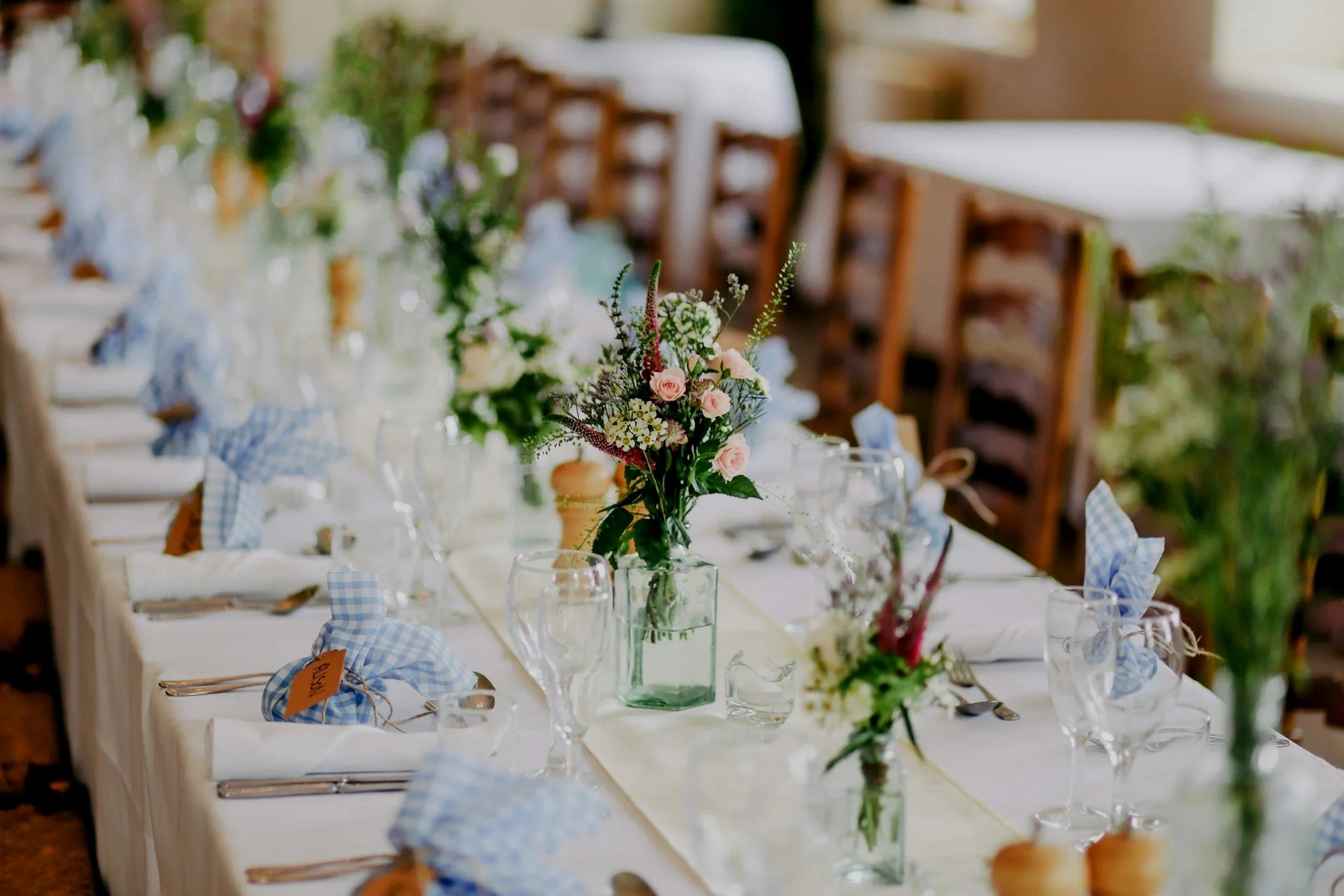 A wedding or formal event reception table decorated with a floral centerpiece, glassware, and blue checkered napkins, in a well-lit, elegant setting.