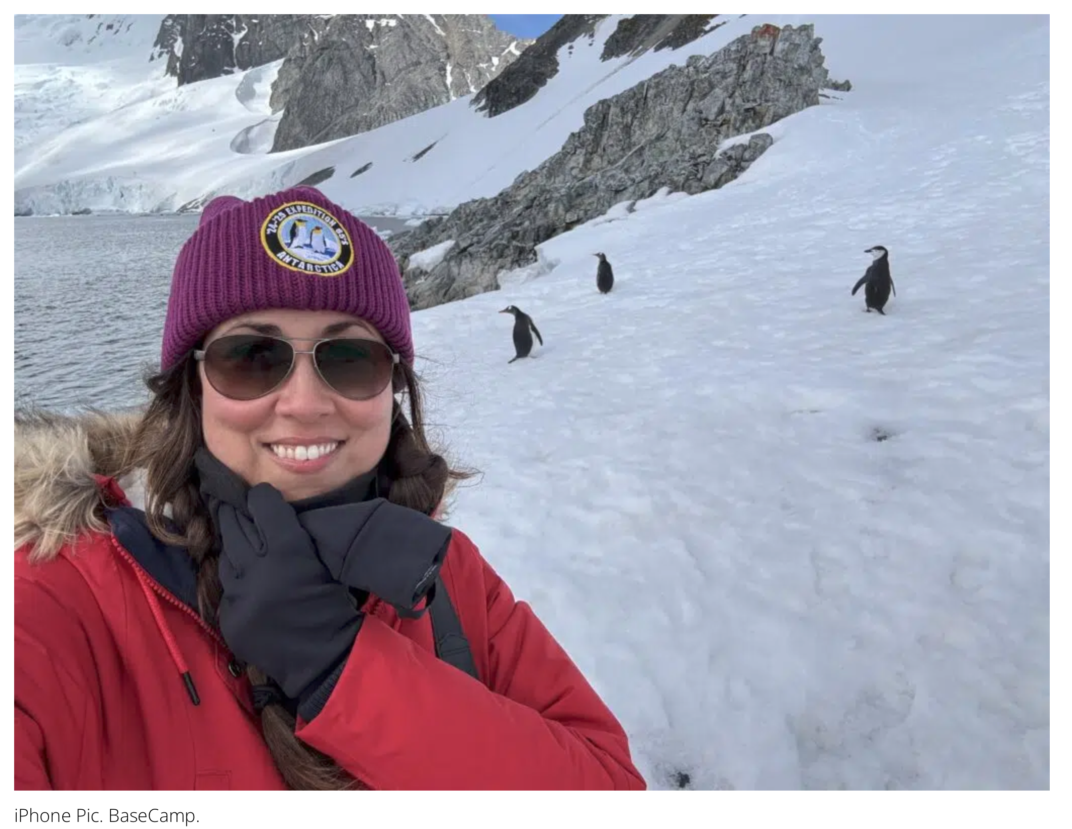 A woman smiling in a red winter jacket and a purple beanie with Penguin Expedition logo, taking a selfie in a snowy mountainous landscape with rocks and three penguins nearby.