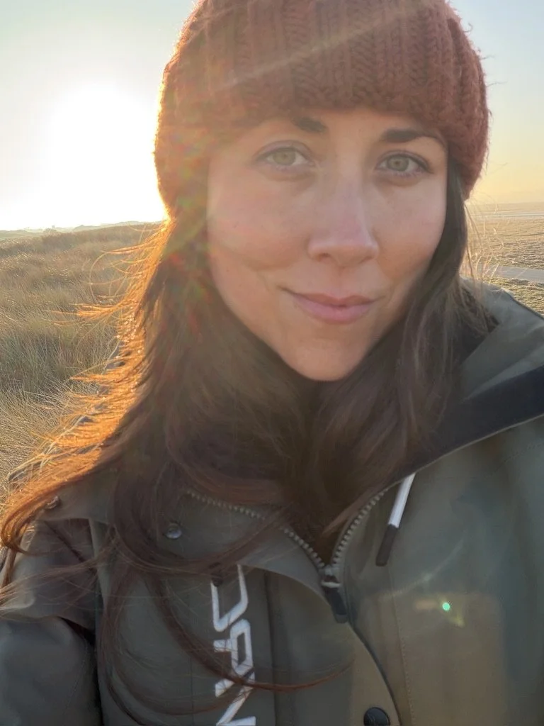 A woman with long brown hair wearing a red knit hat and a gray jacket, standing outdoors during sunset in a grassy area.
