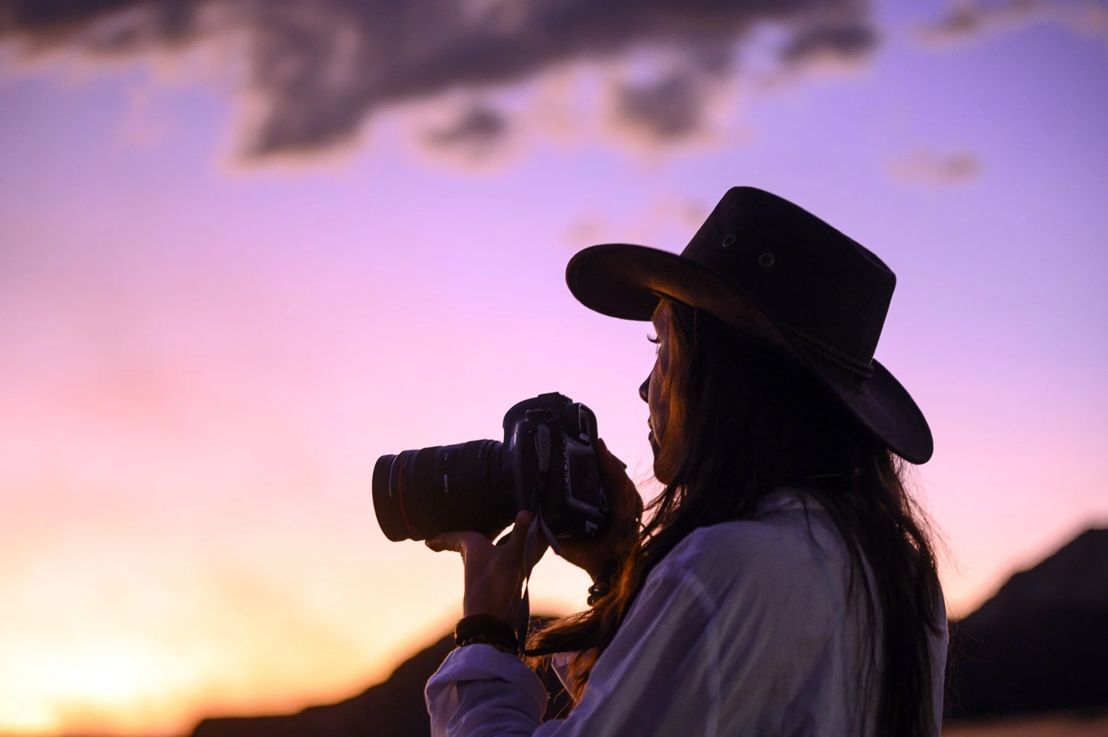 A person wearing a wide-brimmed hat and white jacket is holding a camera, capturing a scene during sunset or twilight with a colorful sky in the background.