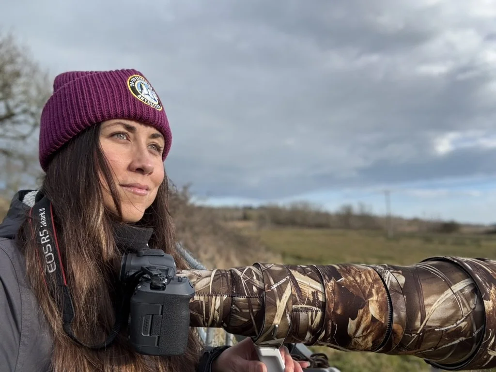 Woman with long brown hair wearing a purple knit cap and black jacket, holding a camera with a camouflage lens cover, standing outdoors on a cloudy day.