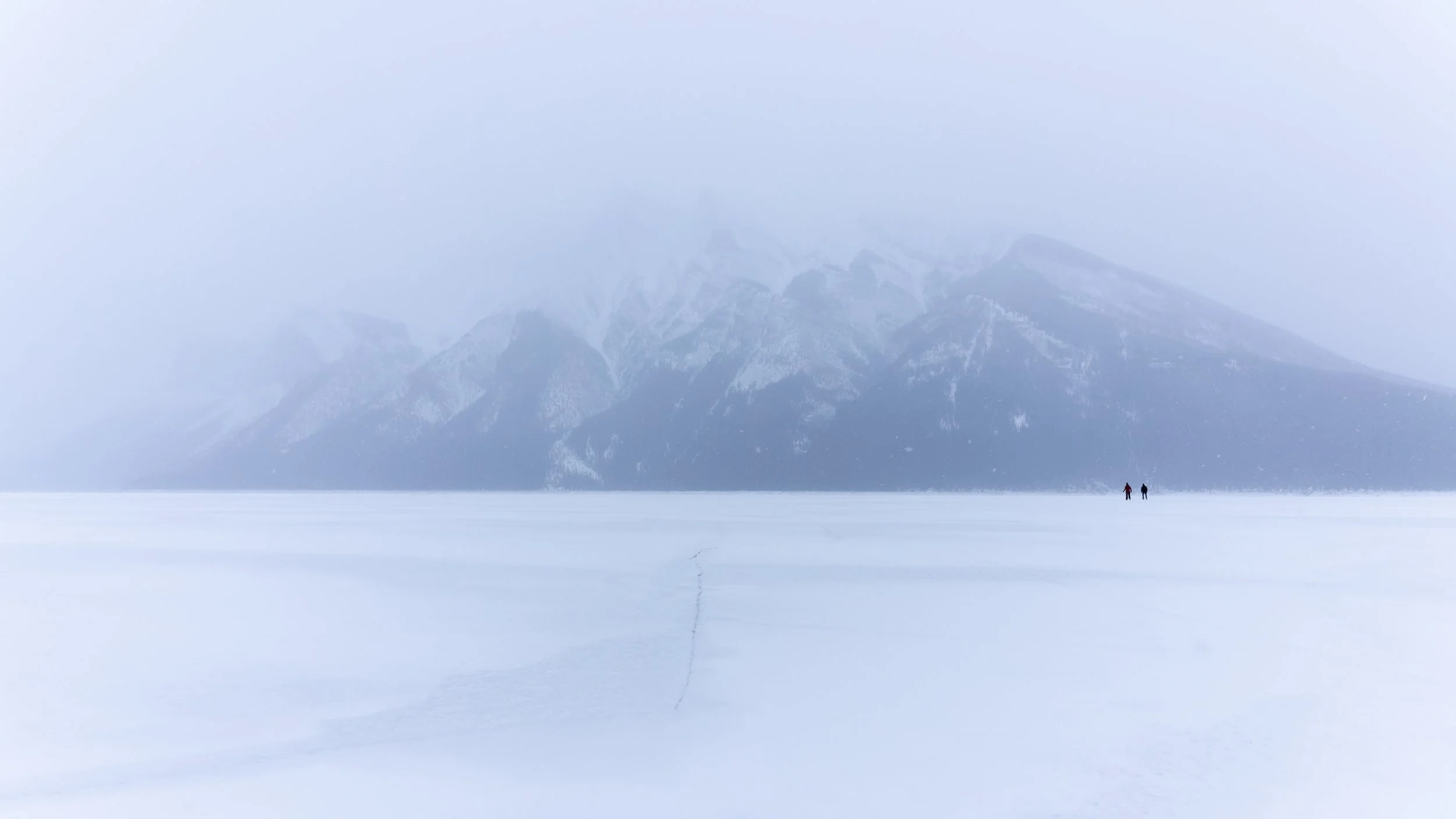 A vast frozen landscape with two tiny figures walking on snow, mountains shrouded in fog in the background.