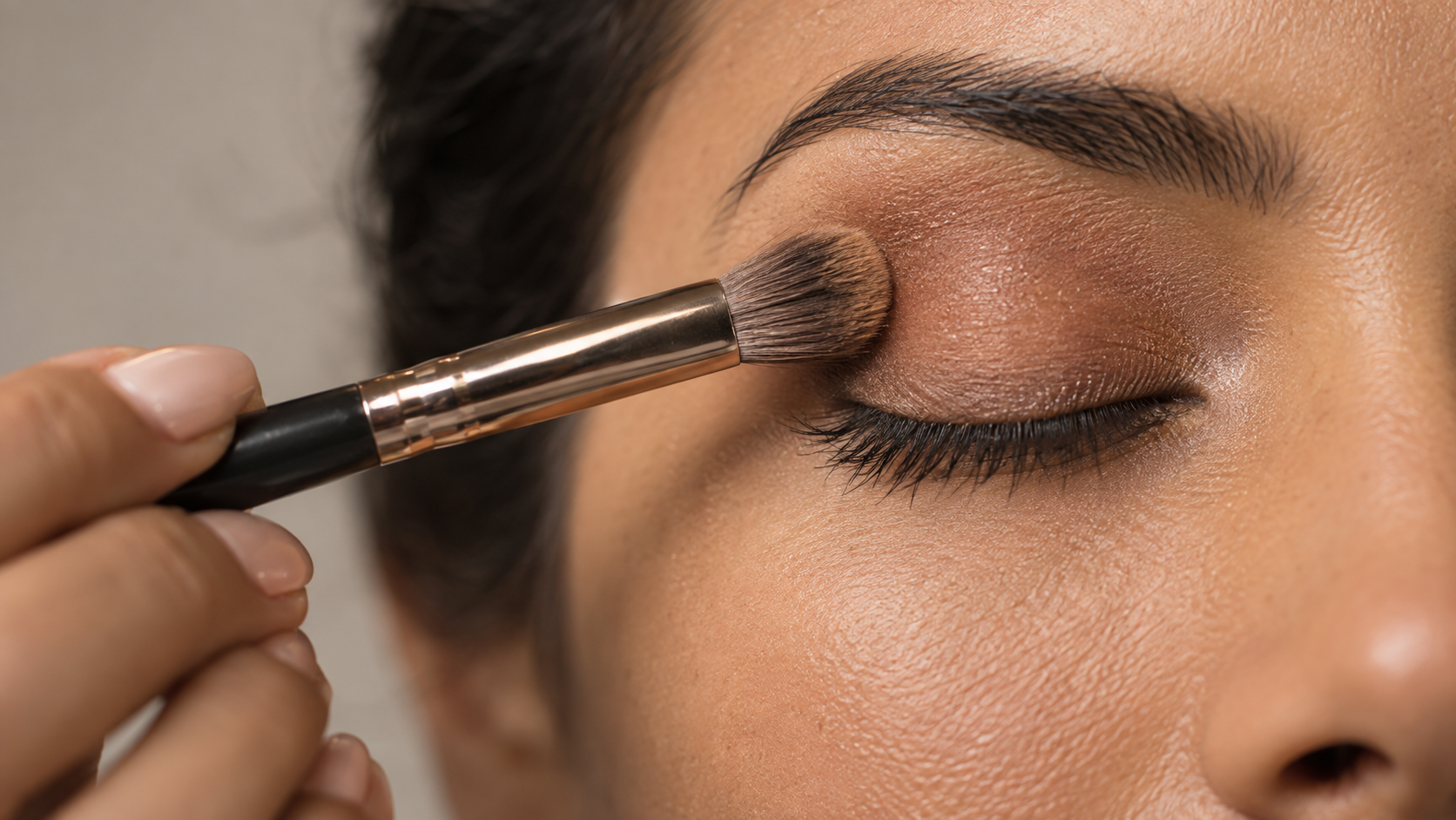 Close-up of a person with medium skin tone having makeup applied to their eyelid using a makeup brush.