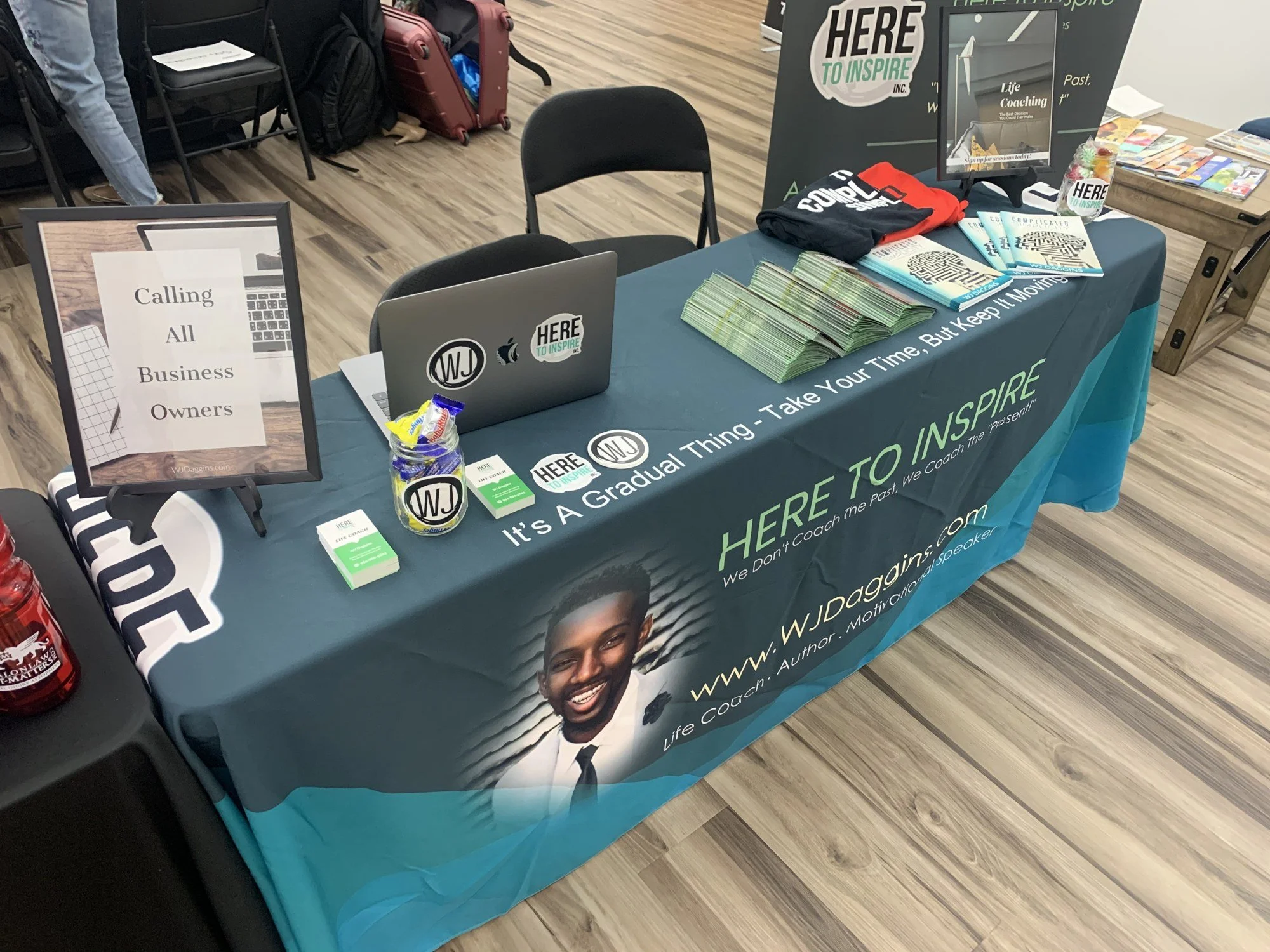 WJ Daggins branded event table with a Here To Inspire banner, business cards and copies of his book, next to a Calling All Business Owners sign