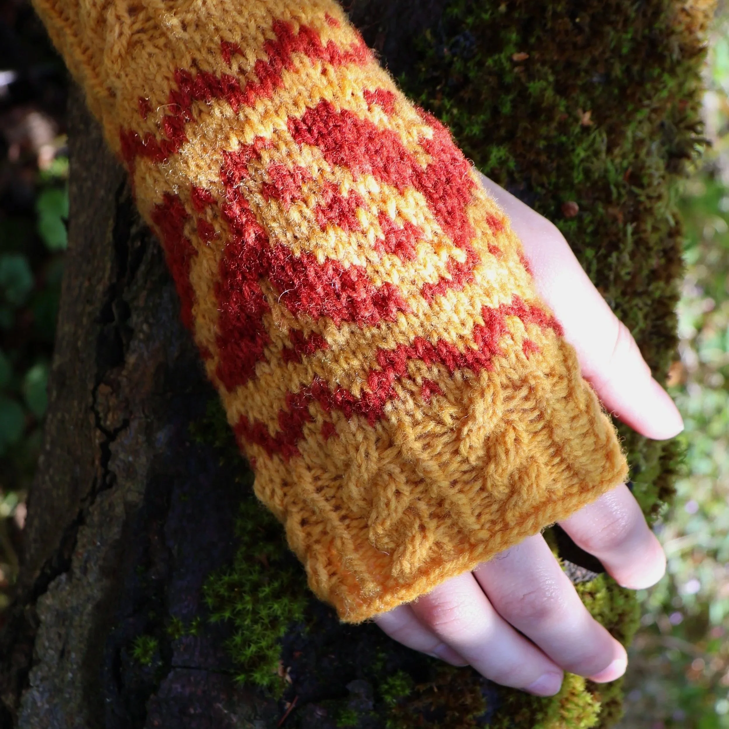 Close-up of a hand with fingers on green moss, wearing a yellow knitted glove with red and orange patterns. Pattern - Kindred Roots for Rooted Textiles.
