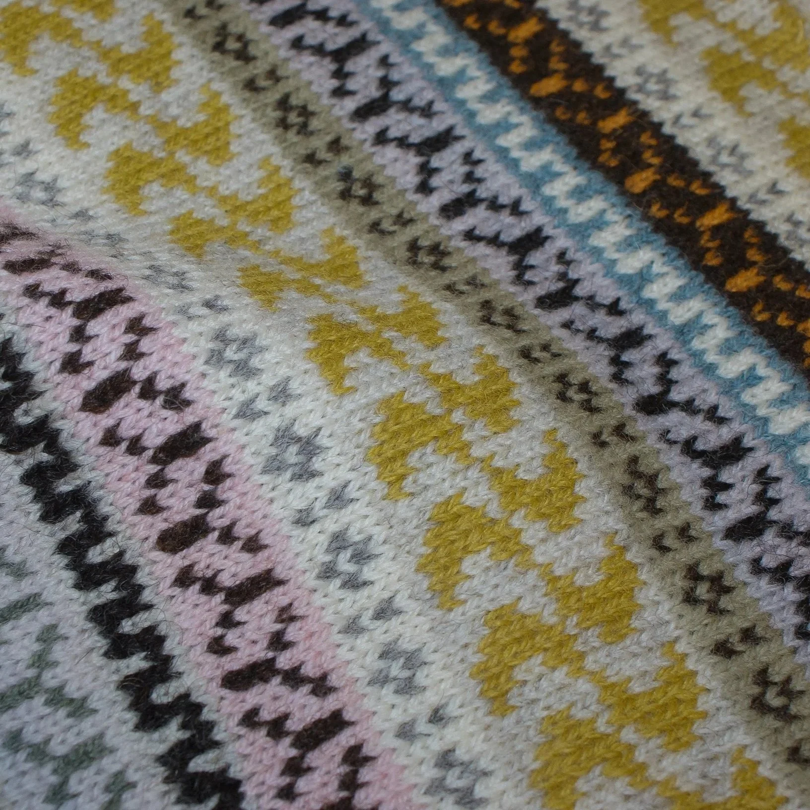 Close-up of a colourful, patterned knitted Fair Isle textile with zigzag and diamond designs in yellow, pink, black, white, blue, and green, inspired by teasels.