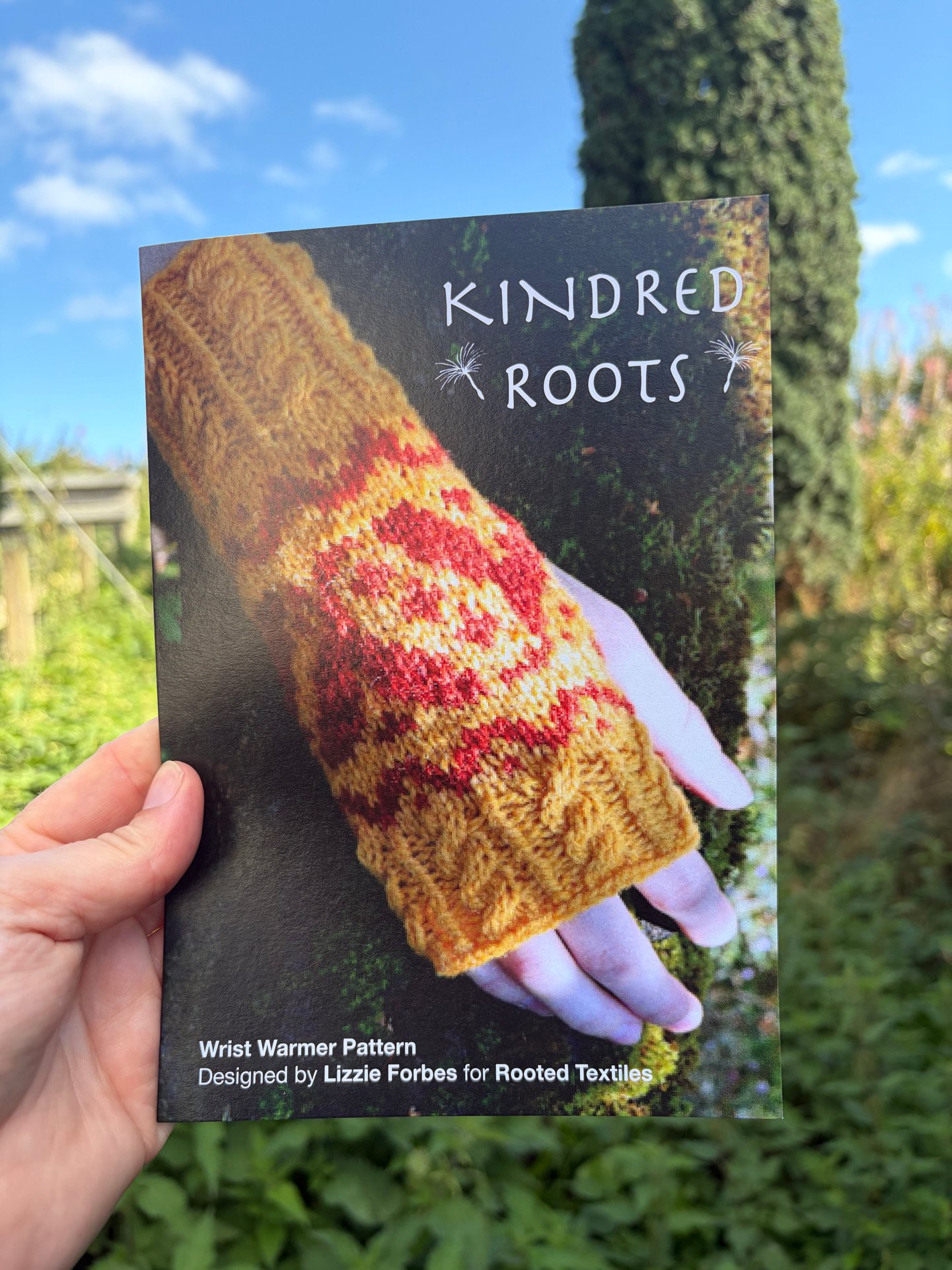 A hand holding a brochure or pattern sheet with a colorful knitted wrist warmer on the cover. The cover reads "KINDRED ROOTS" with dandelion illustrations and states "Wrist Warmer Pattern Designed by Lizzie Forbes for Rooted Textiles." The background is outdoors with green plants and a blue sky with some clouds.
