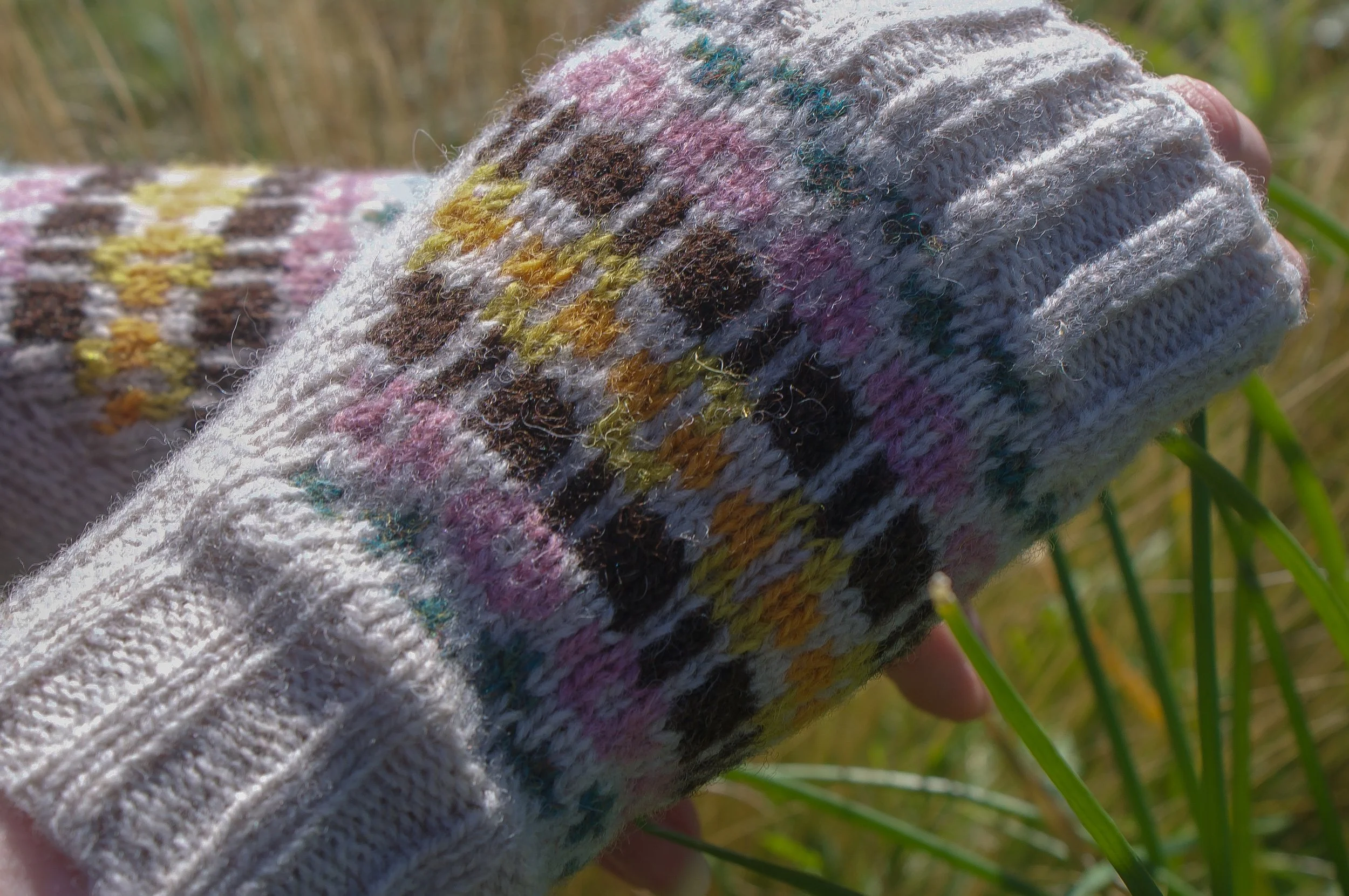 Close-up of a hand wearing a colorful, patterned knit glove holding green grass outdoors.