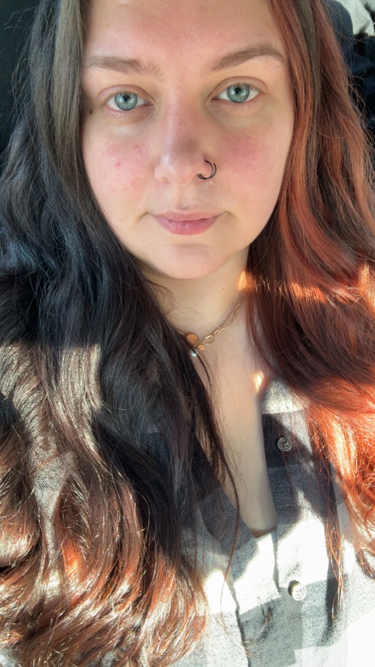 A close-up of a woman with long, wavy dark and red hair, wearing a septum ring, a thin gold necklace, and a plaid shirt, looking directly at the camera with blue eyes.