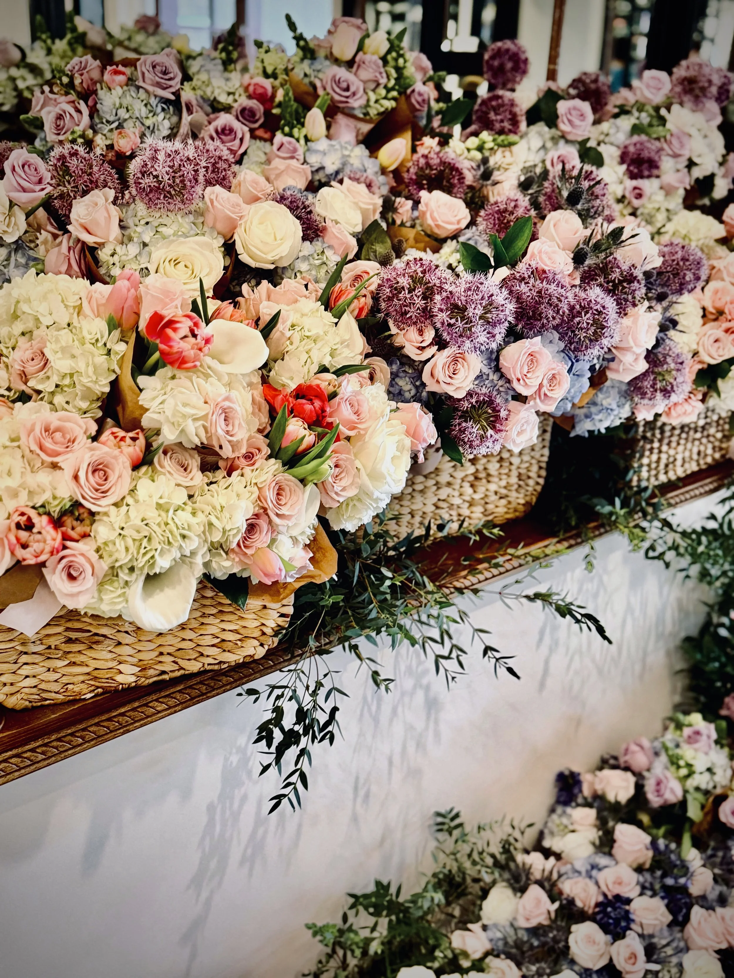 A display of various pink, white, and purple flowers arranged in baskets and on a table, with some greenery.
