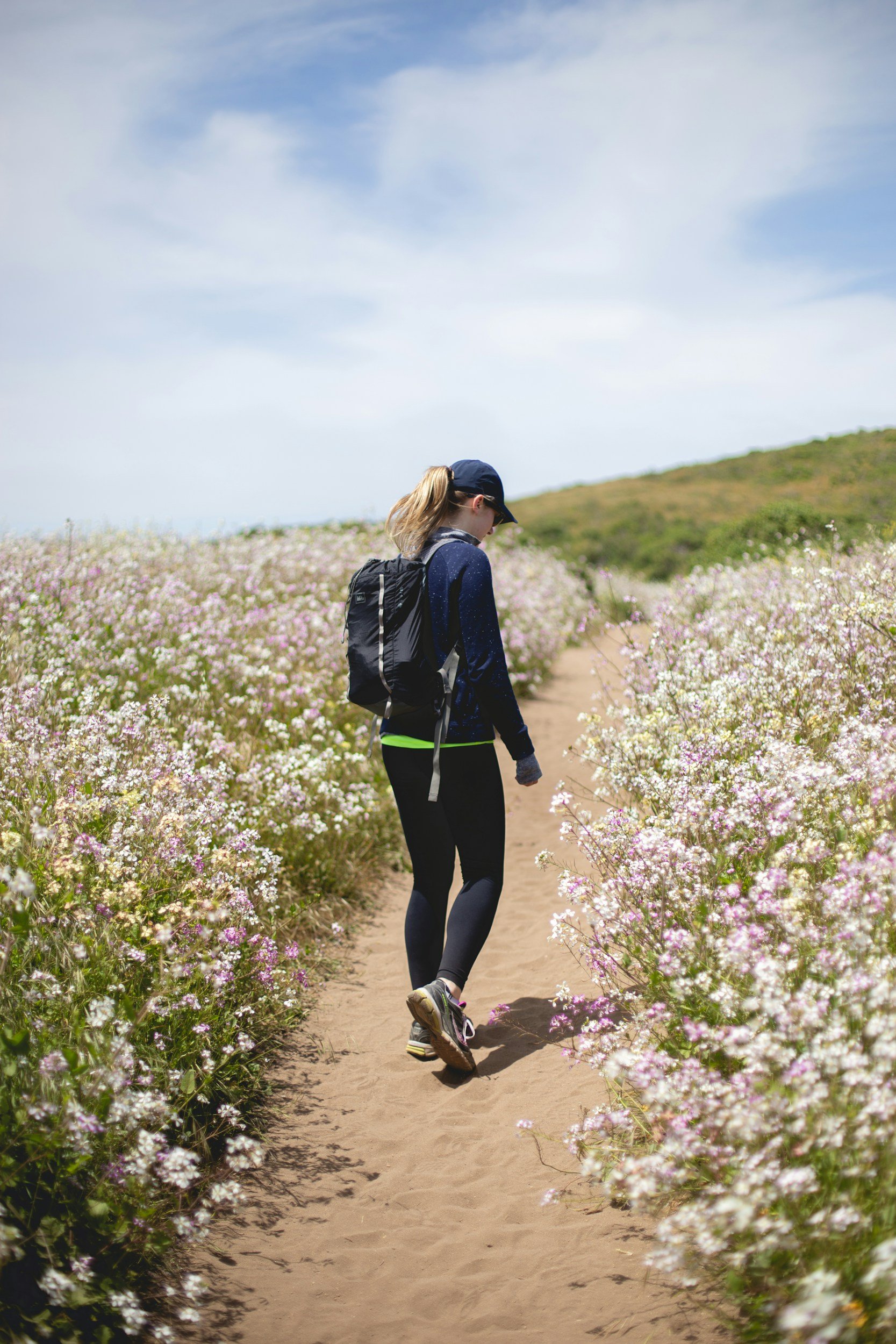 A woman wearing a navy cap, dark jacket, and leggings with a backpack, walking along a dirt trail through a field of pink and white wildflowers under a partly cloudy sky.