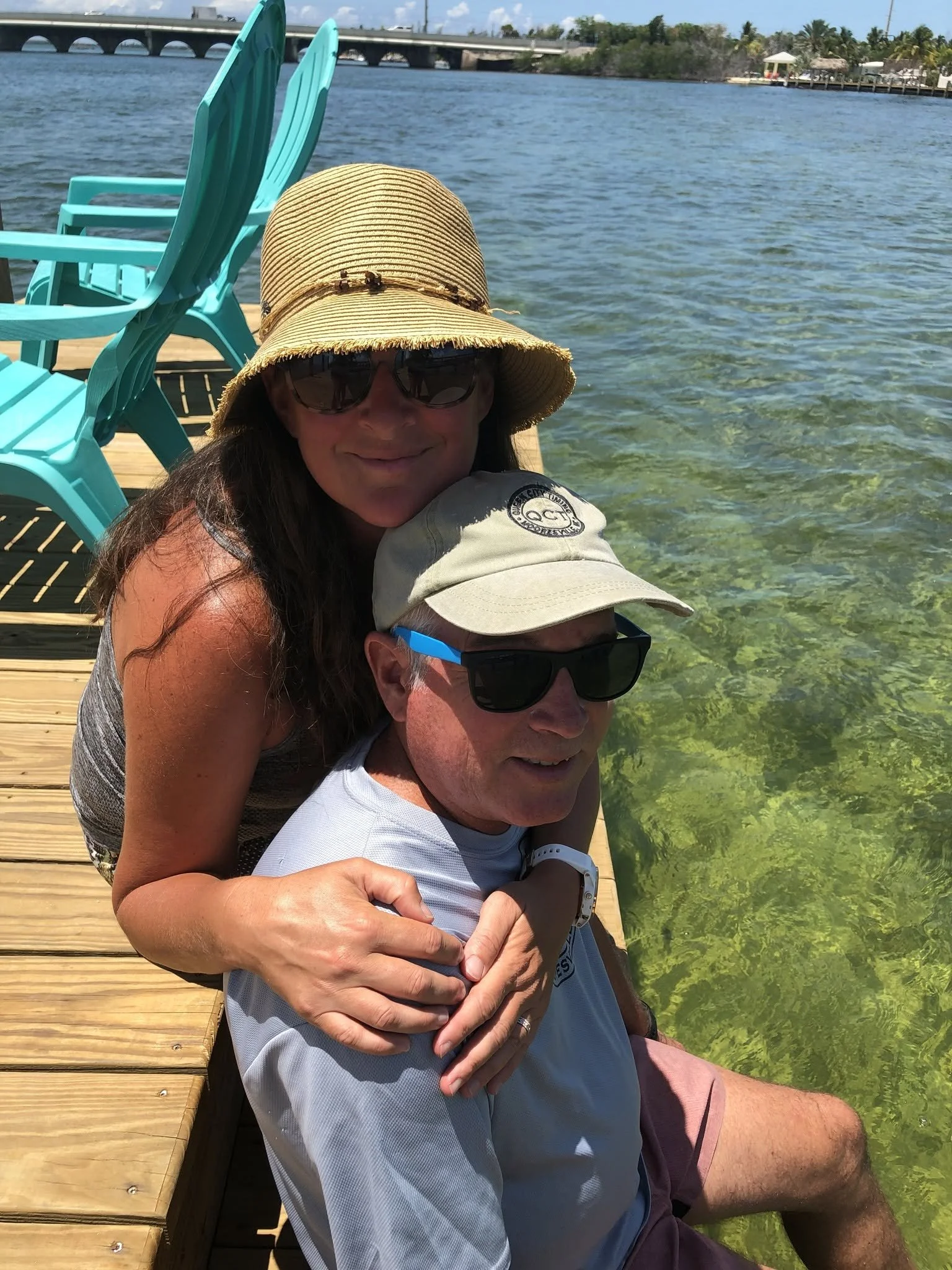 A woman and a man sitting on a dock by a river or lake, with the woman wearing a straw hat and sunglasses, and the man wearing sunglasses and a cap. The background includes turquoise chairs, water, a bridge, and some houses or buildings.