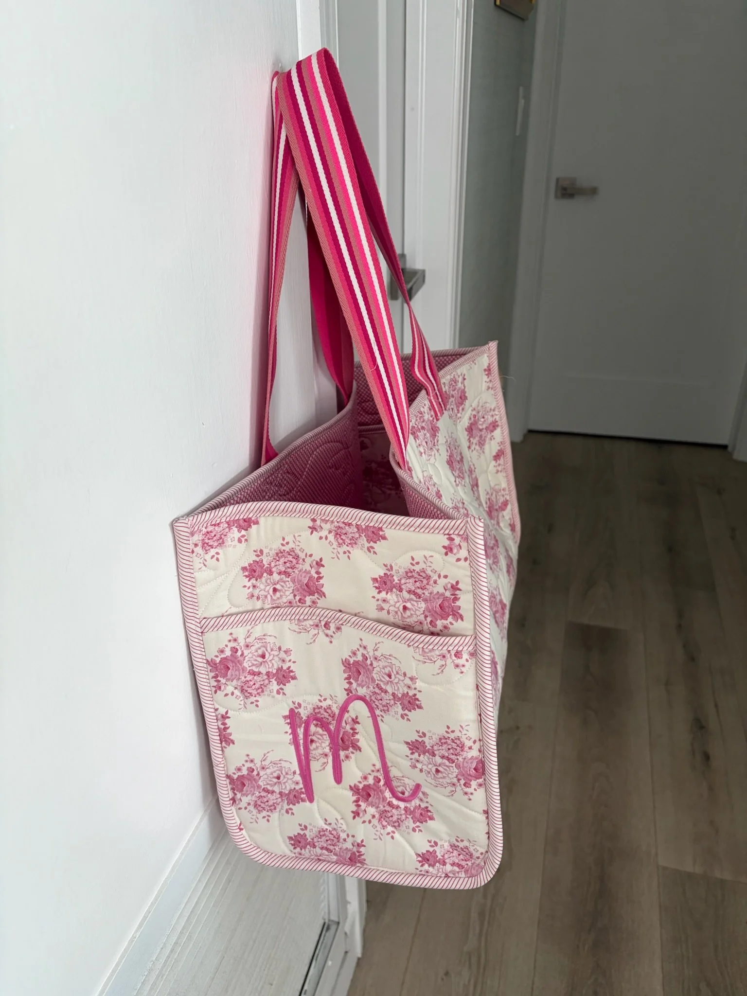A pink and white floral tote bag. The bag has pink handles and the initial 'M' written on it in pink.