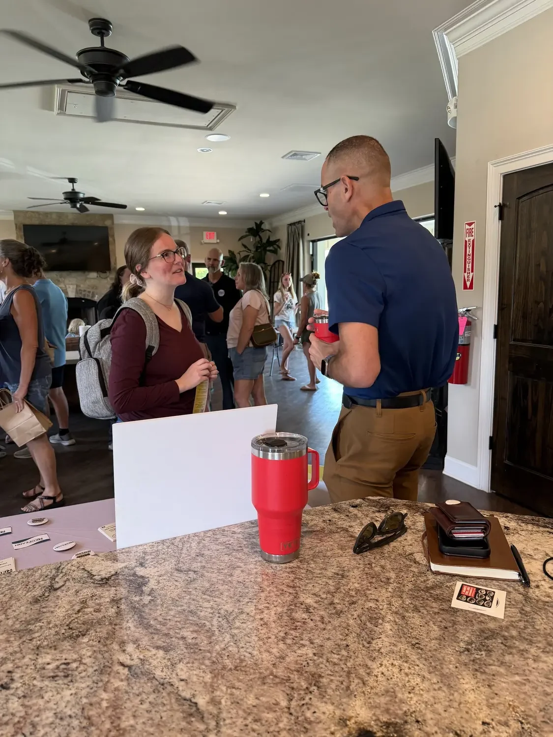 A woman with glasses and a backpack engaging in conversation with a man holding a red tumbler in a room with several people, a television, and a stone fireplace visible in the background.