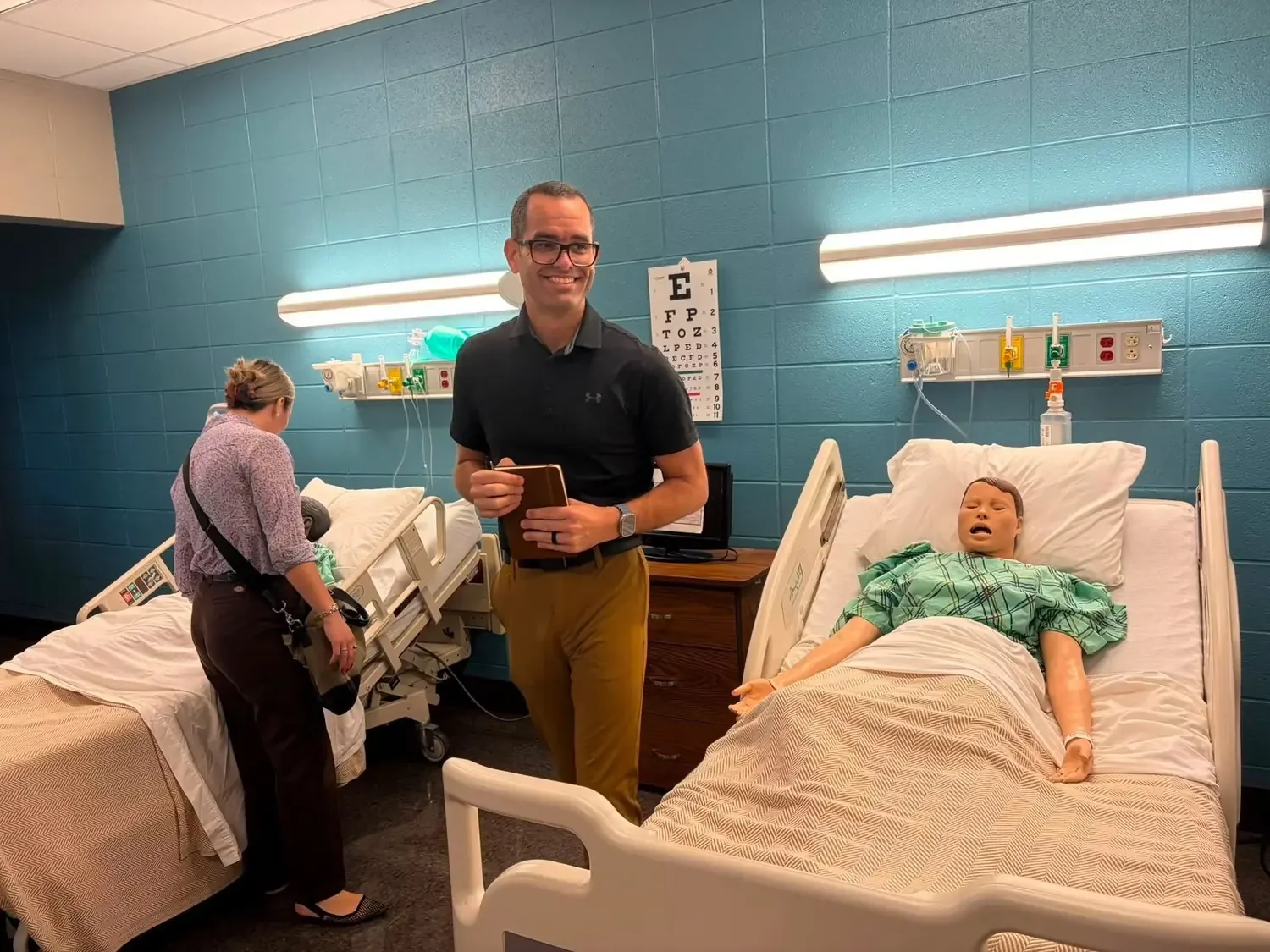 A scene in a hospital room with two beds, one occupied by a medical training mannequin with a bandage on its arm, the other unoccupied, and two healthcare professionals preparing equipment. A smiling man with glasses is standing in the middle, holdin