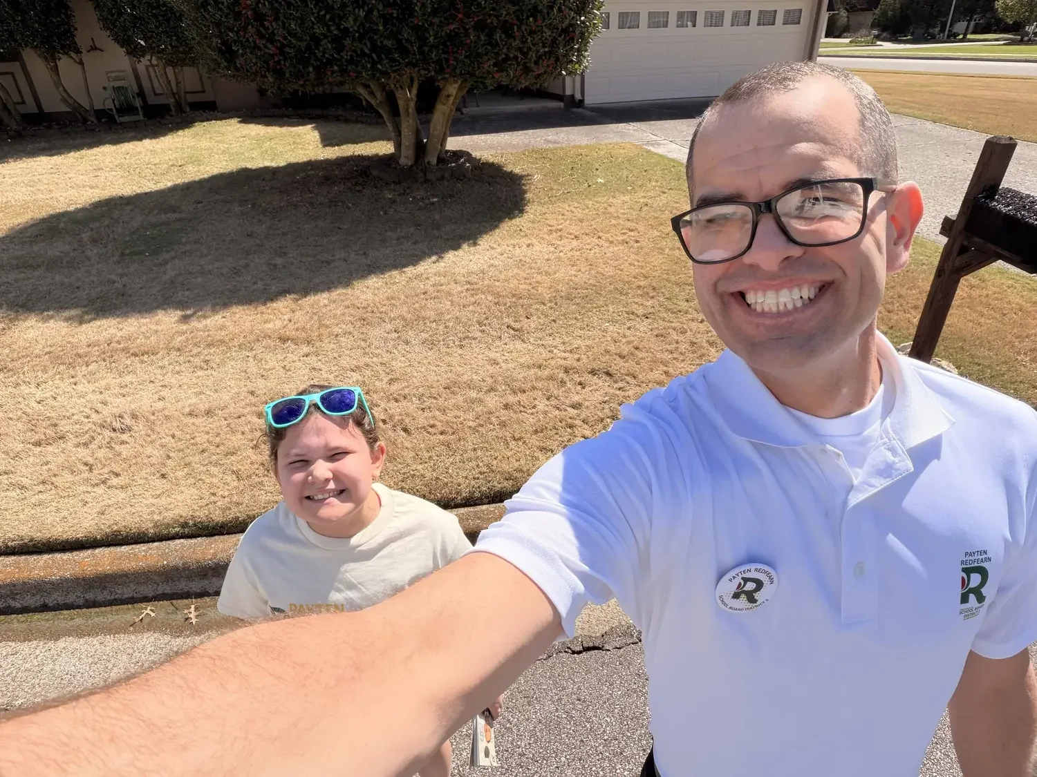 A man taking a selfie with a young girl outdoors on a sunny day. Both are smiling, the girl wears sunglasses on her head and a white shirt, the man wears glasses and a white polo shirt with logo patches.
