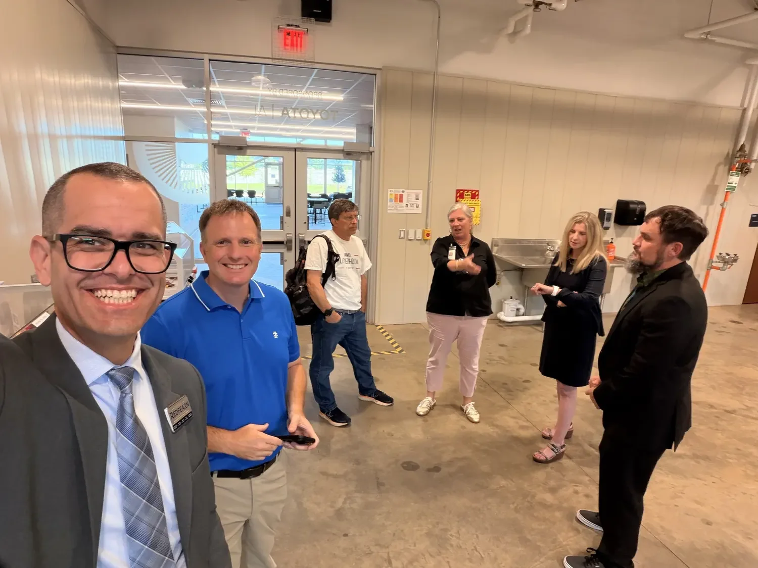 Six people standing and talking in an industrial or cafeteria setting. Two men in the foreground are smiling and taking a selfie. The man on the left wears a suit, tie, glasses, and a name tag. The man next to him is in a blue polo shirt. The other f
