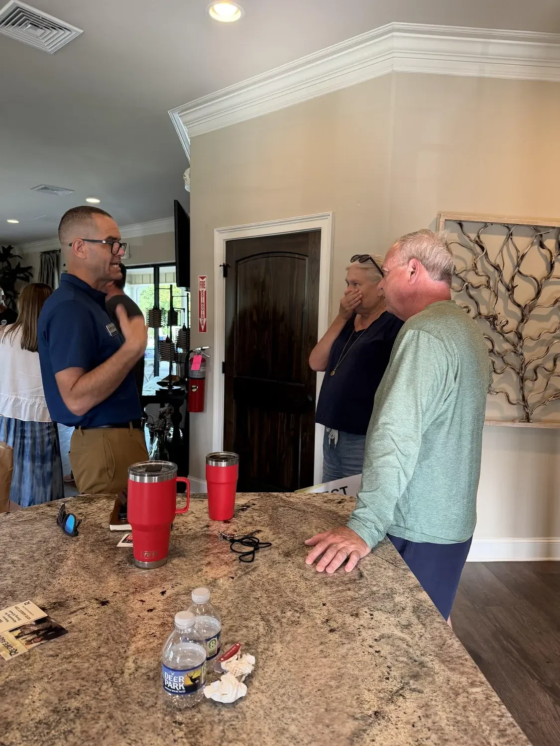 A man in a navy blue shirt is talking to two women at a kitchen counter. The women appear to be listening intently; one has her hand over her mouth, and the other has her hand on the counter. There are two red tumblers, a pair of sunglasses, water bo