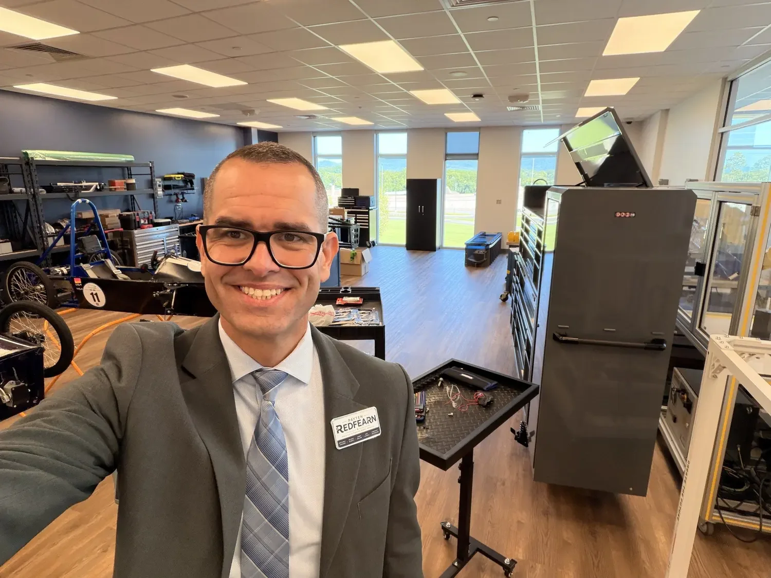 A man in a suit and tie taking a selfie inside a workshop filled with equipment and tools, with large windows and a view of green fields outside.