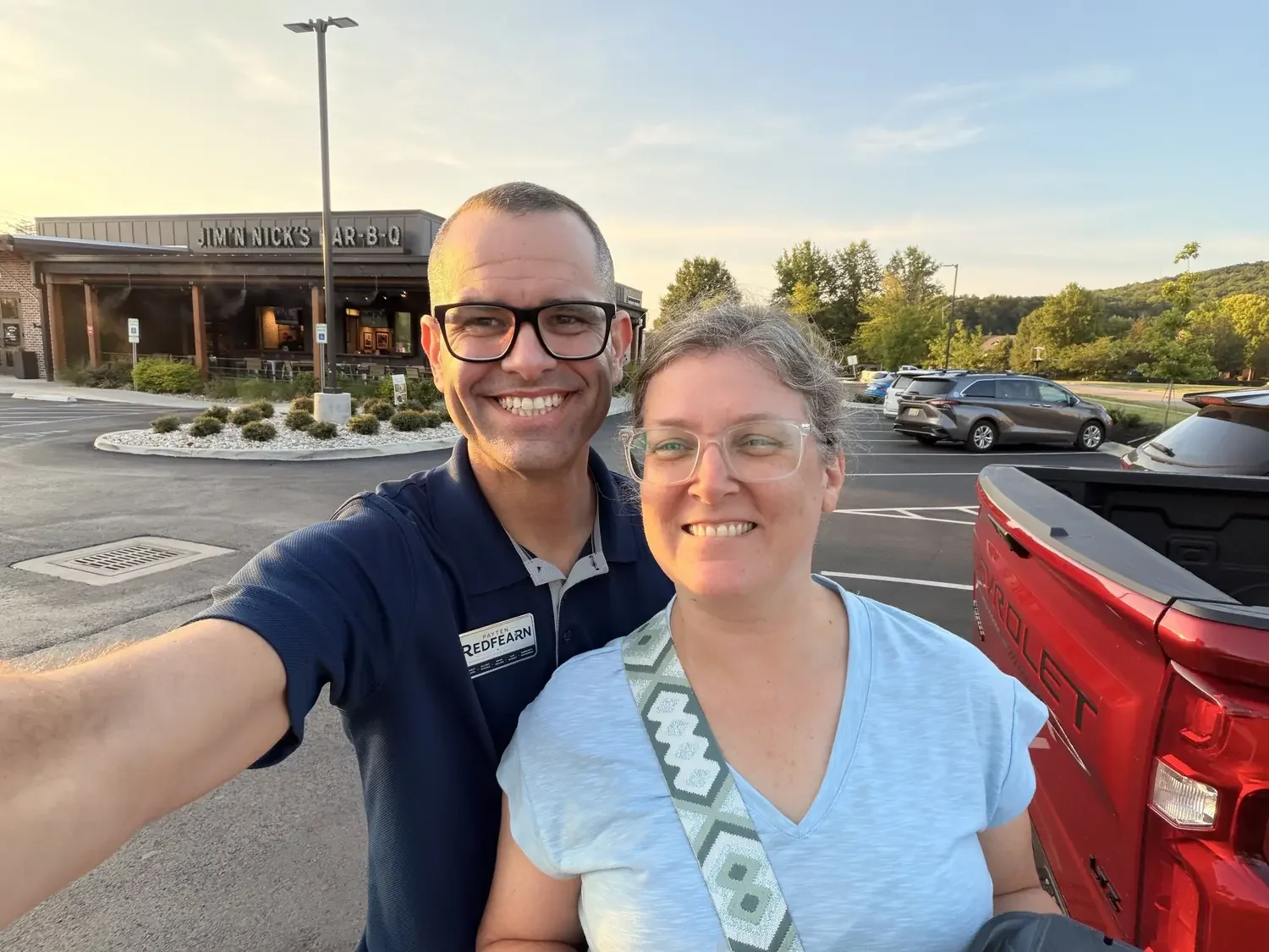 A smiling man and woman taking a selfie in a parking lot during sunset, with a restaurant named Jim N Nick's Bar-B-Q and greenery in the background.