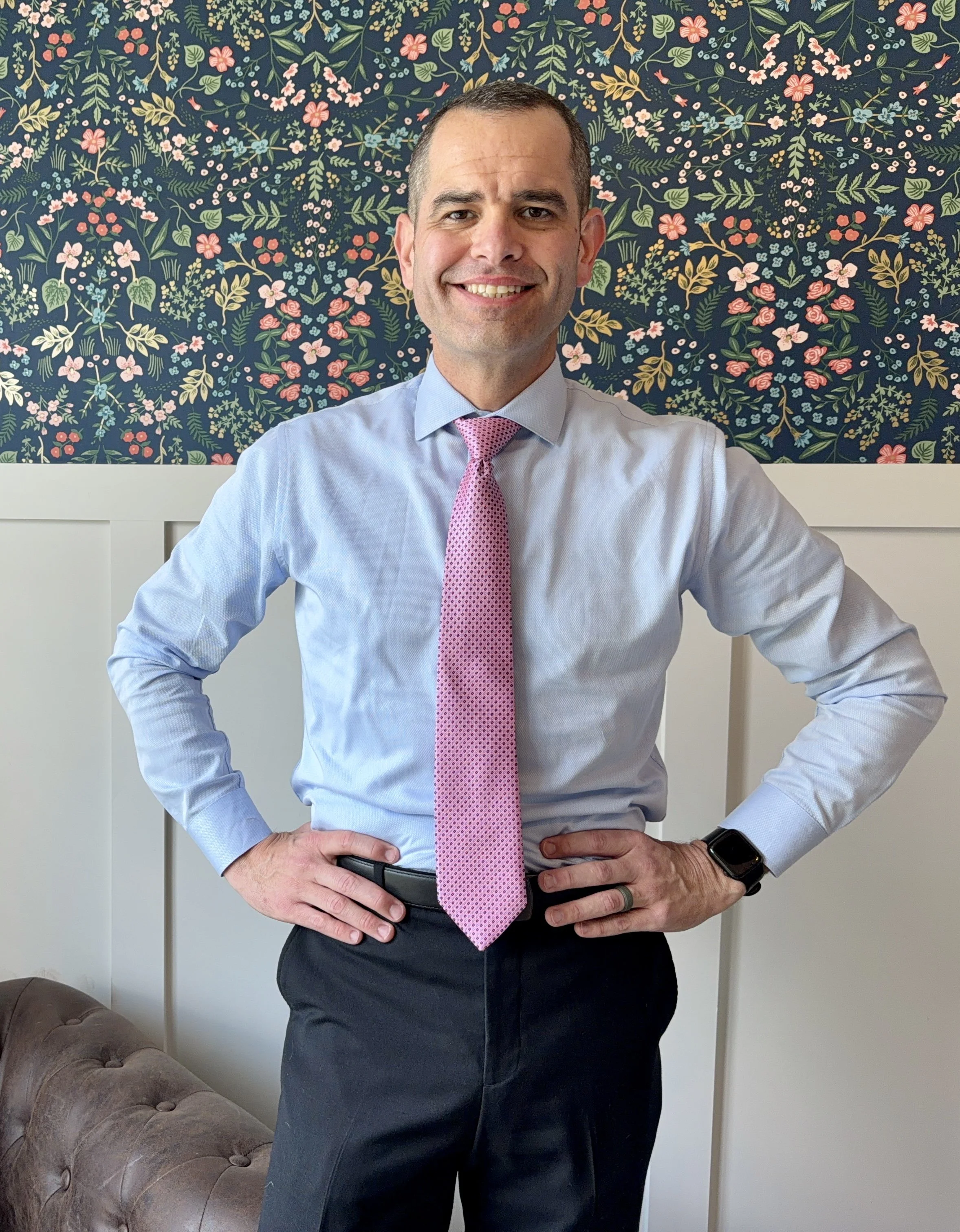 A man in business attire, wearing a light blue shirt, pink tie, and a smartwatch, stands with hands on hips in front of a floral wallpaper background inside a room with a white door and a brown tufted sofa.
