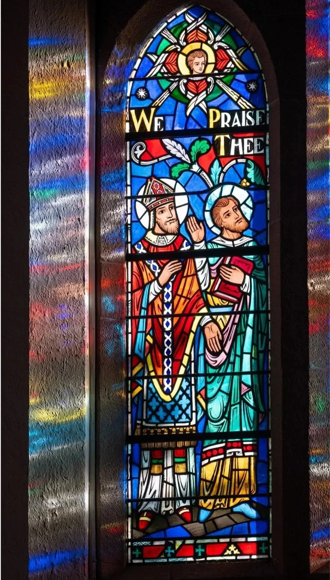 Colorful church stained-glass window depicting Jesus and a bishop beneath the words “We praise thee.”