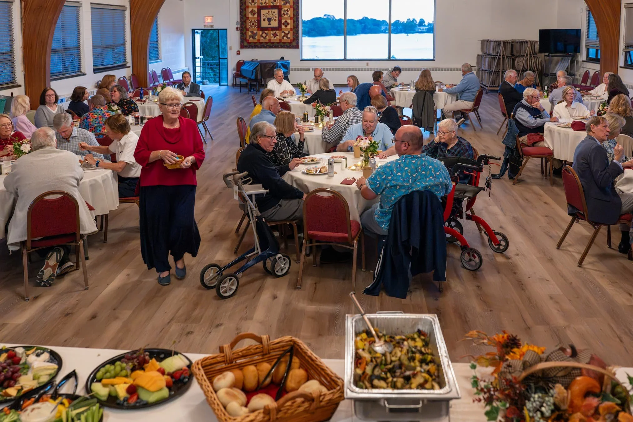 Community luncheon with seniors dining at round tables in a lakeside banquet hall, featuring a buffet of fruit, bread, salad, and roasted vegetables.