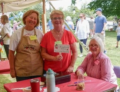 Three women smiling at a community event booth, with a table of refreshments and other attendees in the background